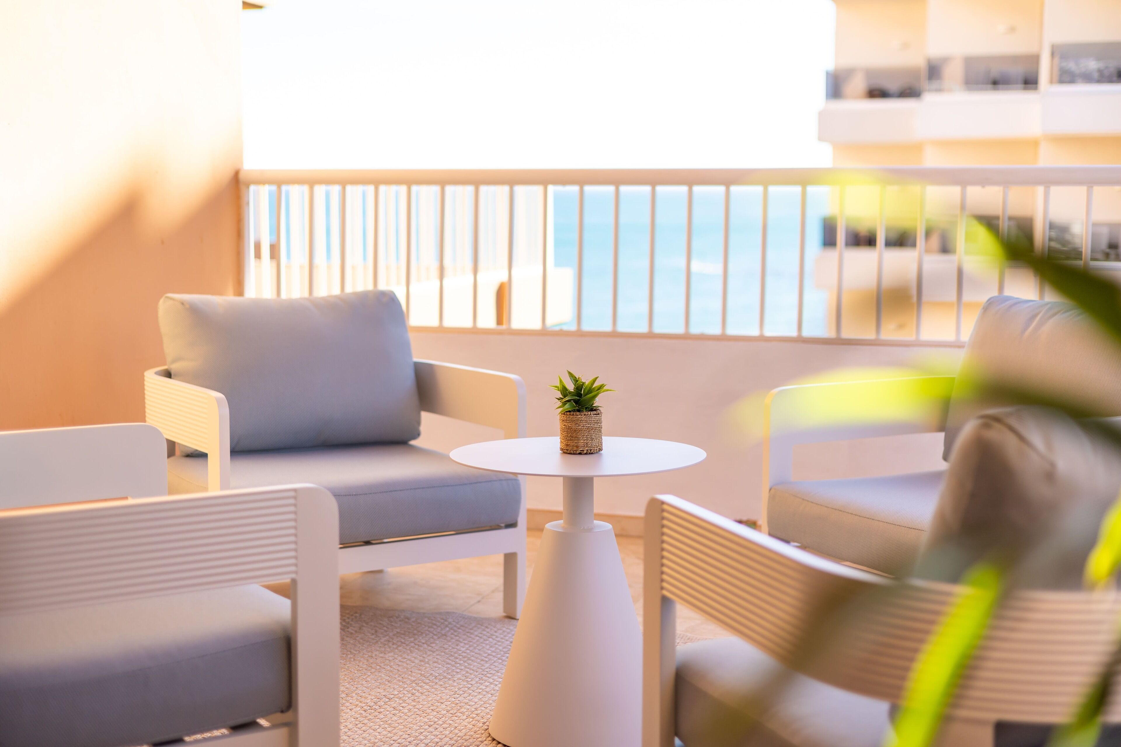 Un área de estar moderna con una mesa redonda y dos sofás, una planta pequeña sobre la mesa, y vista al mar a través de la barandilla del balcón.
