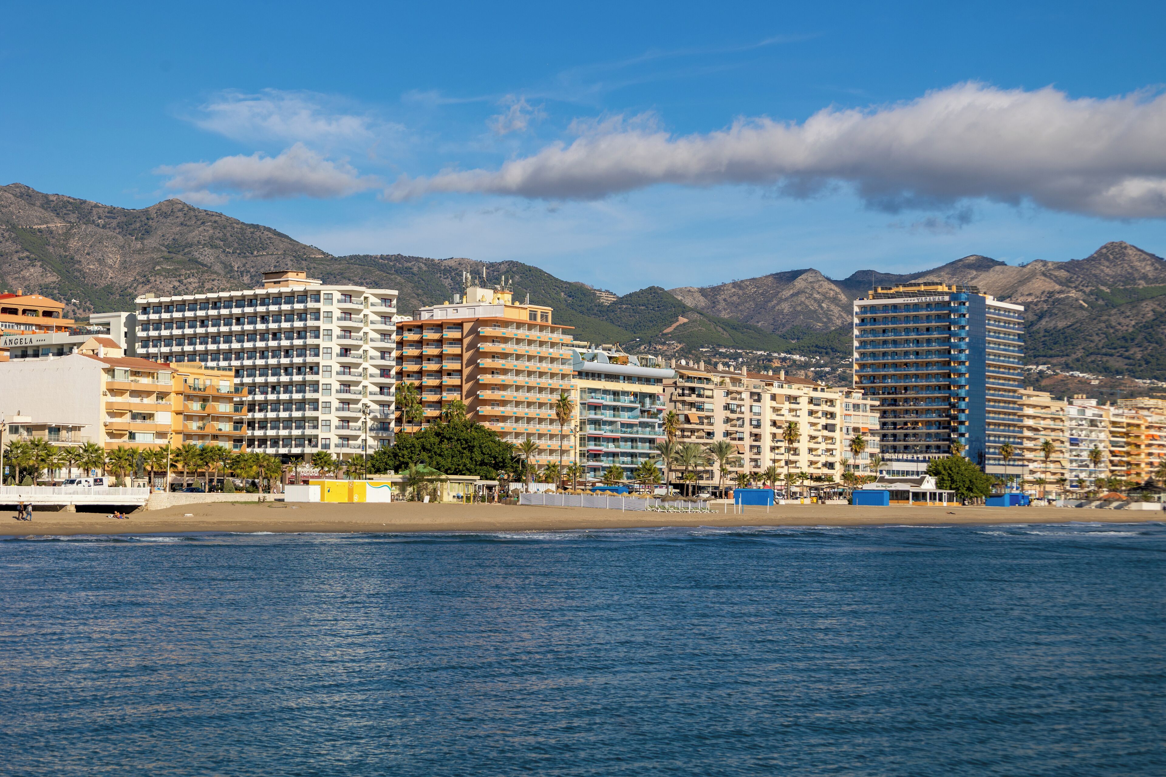 Un paisaje costero con hoteles de varios pisos, una playa y montañas al fondo.
