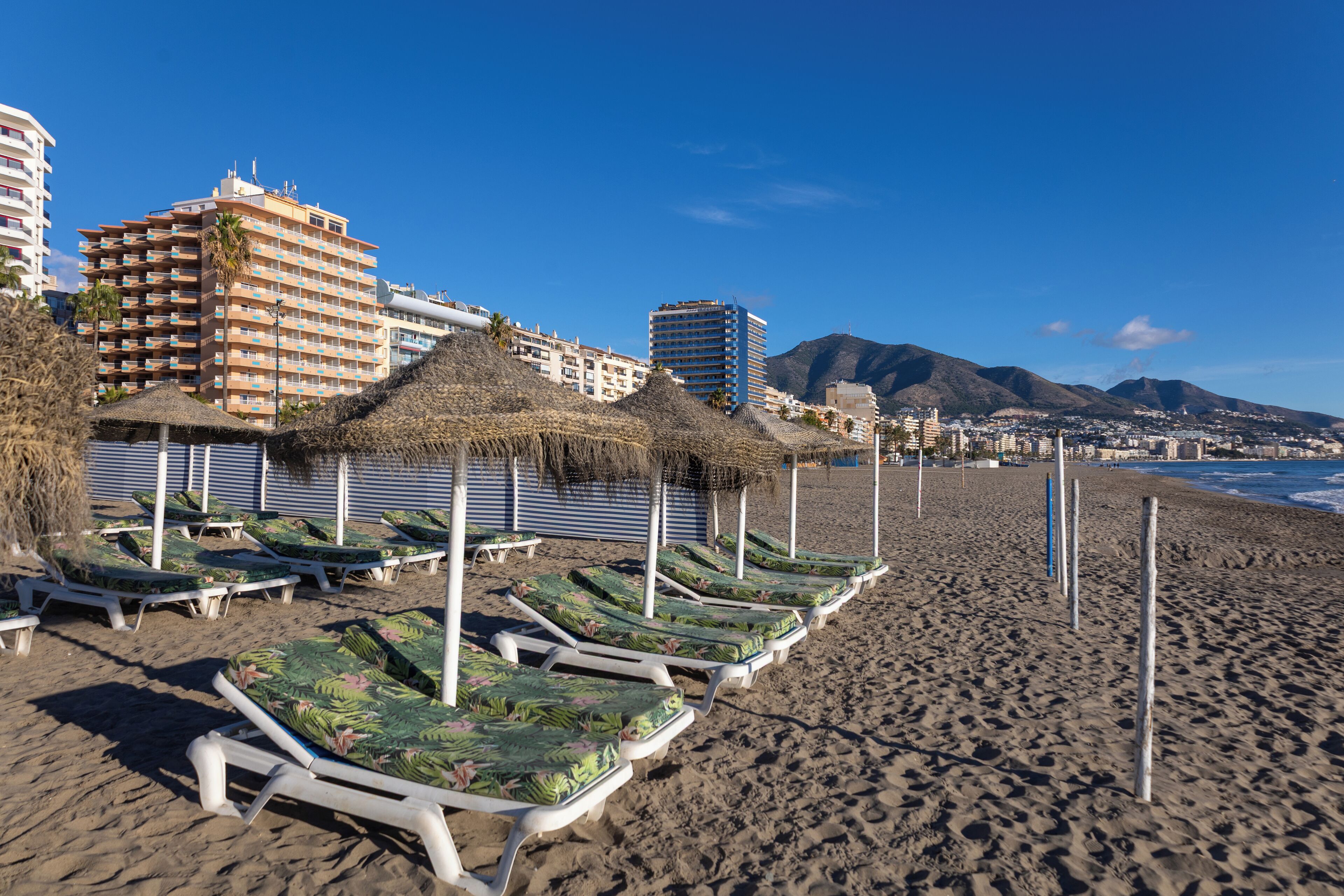 Una playa con filas de tumbonas y sombrillas de paja, un hotel al fondo y una montaña en la distancia.
