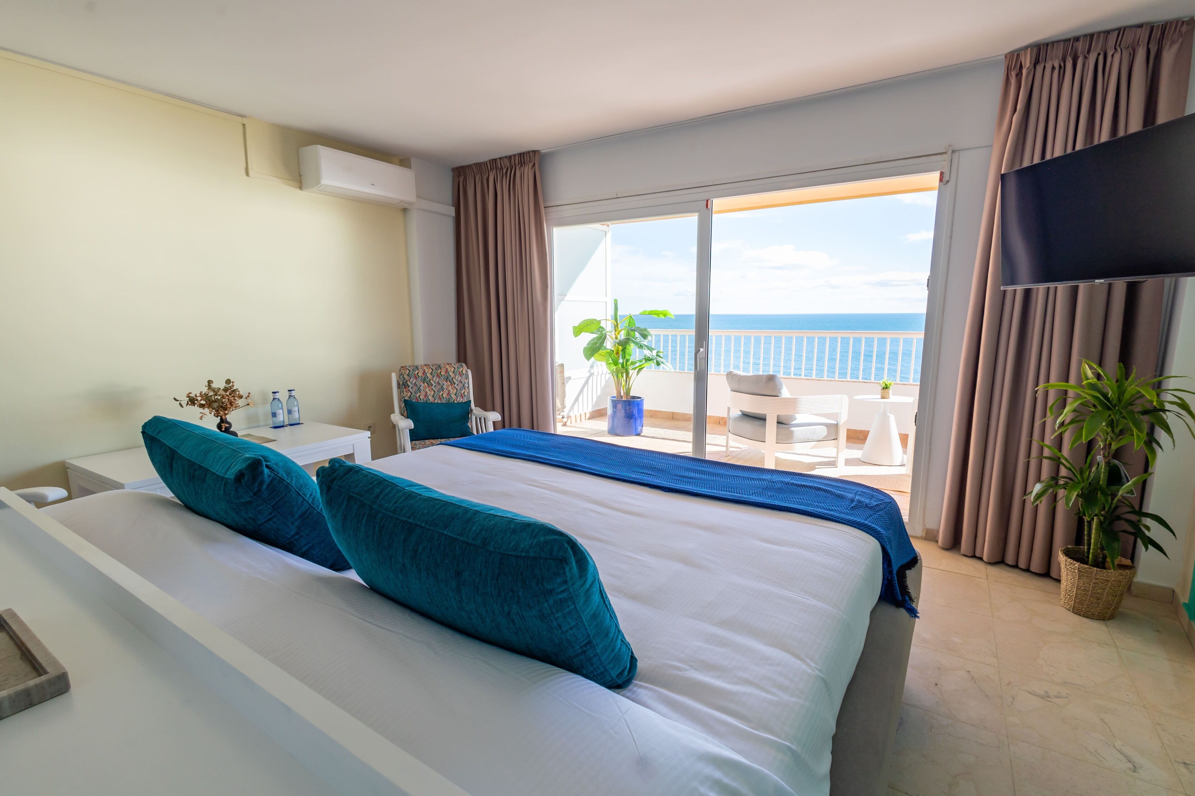 A hotel room with a large bed, a TV, a balcony with a view of the sea, and a potted plant.