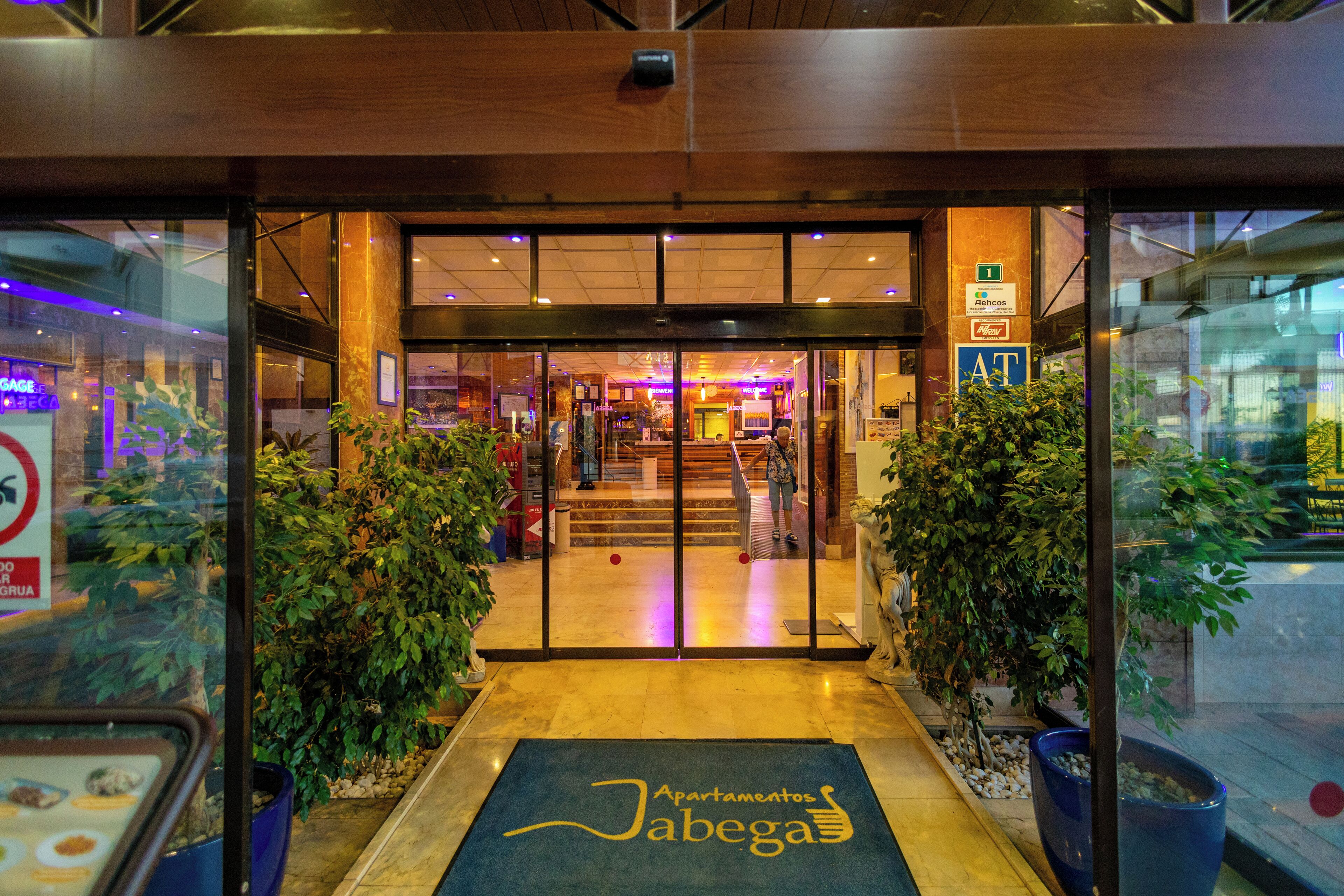 Entrada de un hotel con puertas de vidrio, un mostrador de recepción y un felpudo azul con el nombre del hotel.