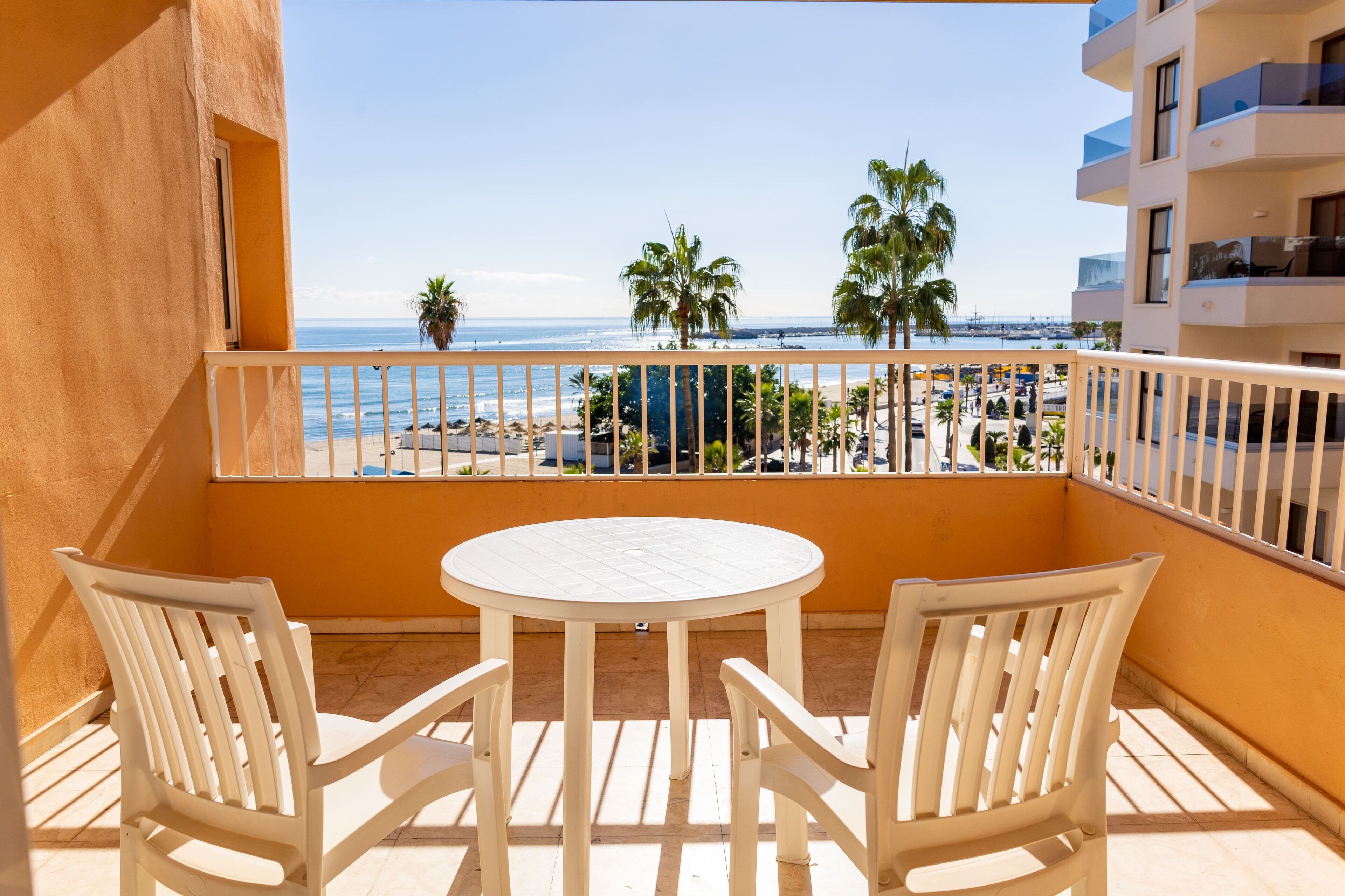 A balcony with a white table and chairs, overlooking a beach with palm trees and a clear sky.