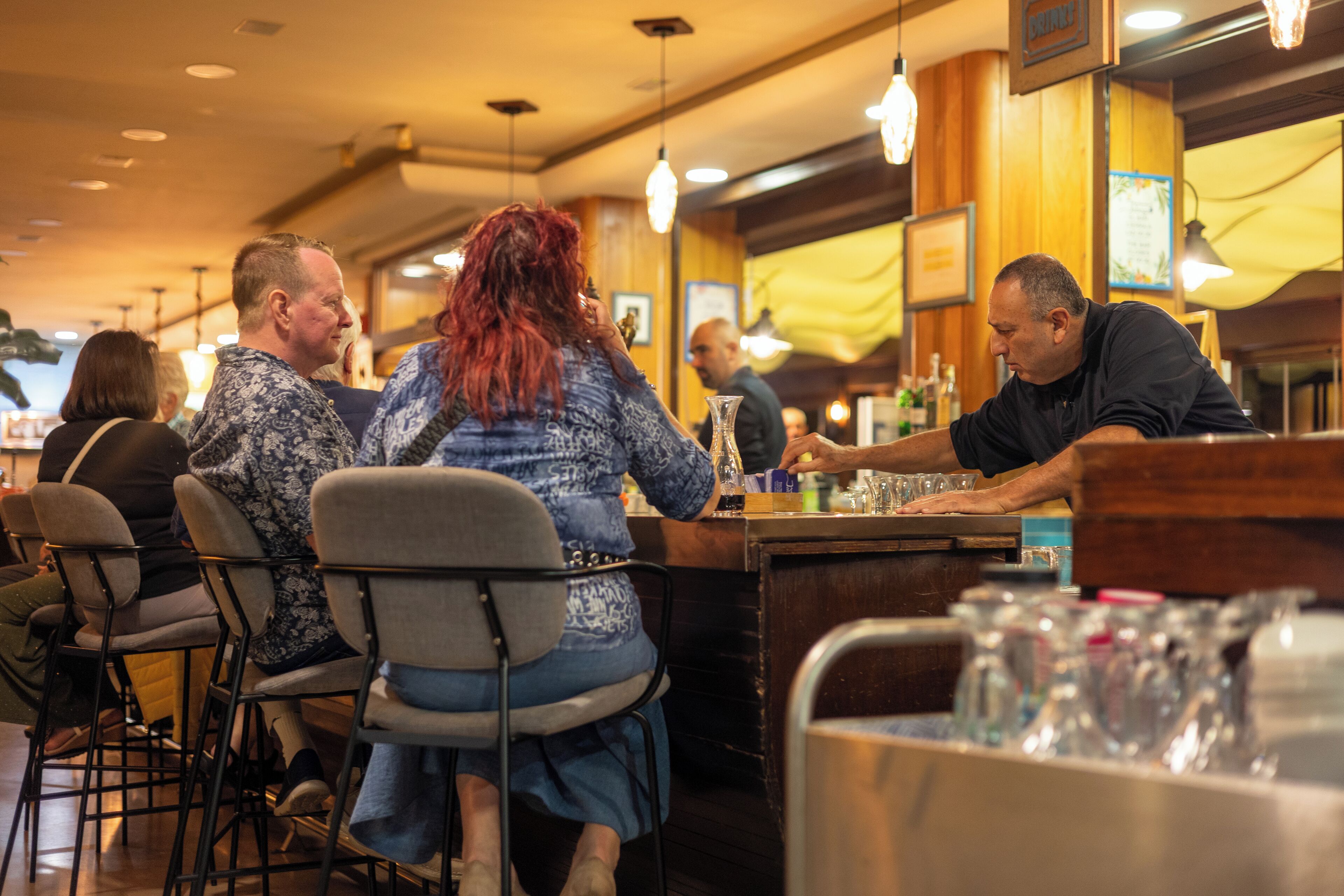 A bar area with patrons seated at tables, a bartender at the counter, and various bar items on the counter.