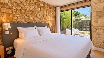 A bedroom with a stone wall, a bed with white linens, two bedside lamps, and a view of an outdoor area with a white chair and umbrella.