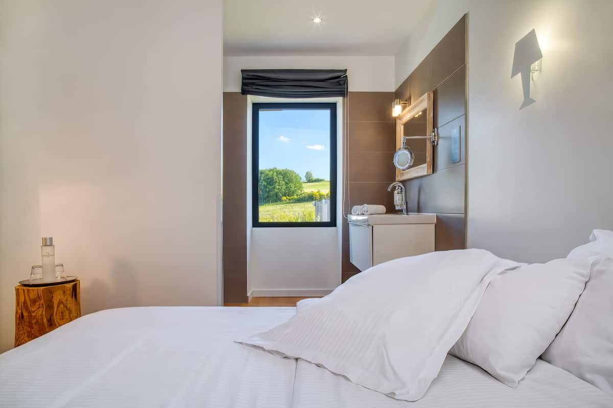 Une chambre à coucher avec un grand lit, une table de chevet sur laquelle se trouvent une bouteille et des verres, un miroir et une vue sur un paysage verdoyant par la fenêtre.