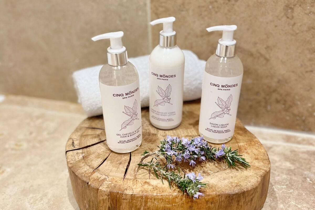 Trois flacons de lotion pour le corps de la gamme Cinq Mondes Spa Paris, posés sur une surface en bois, avec une serviette et des fleurs.
