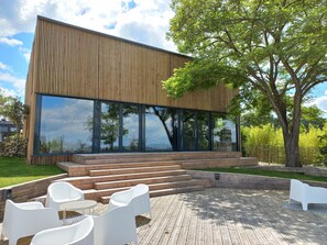 A modern building with large glass windows, a wooden exterior, and a deck with white chairs and a table.