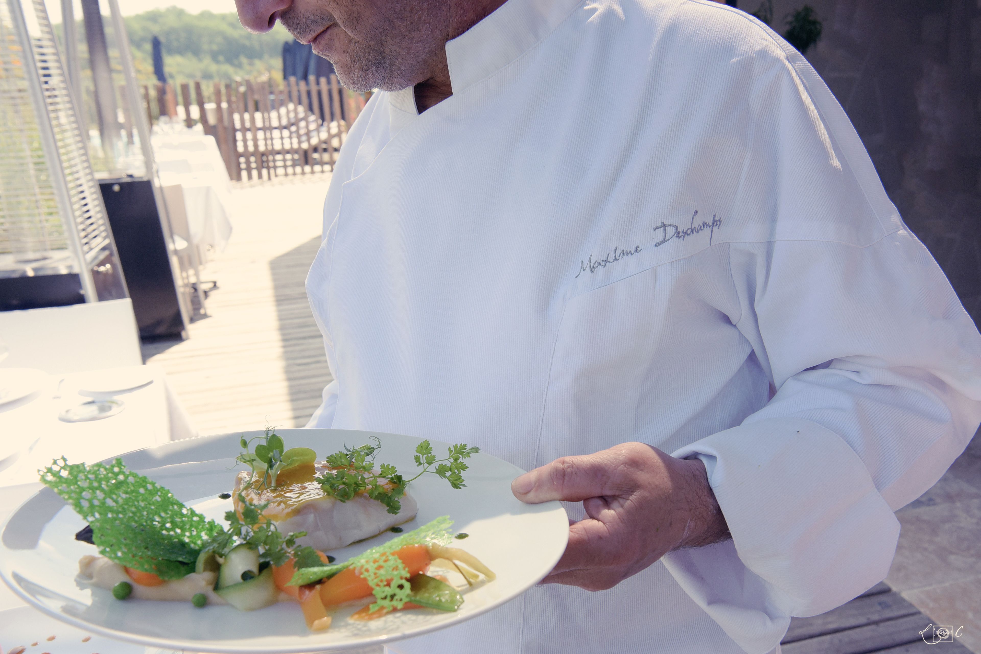 Um chef segurando um prato de comida, com vista para um deck de madeira e mesas ao ar livre.