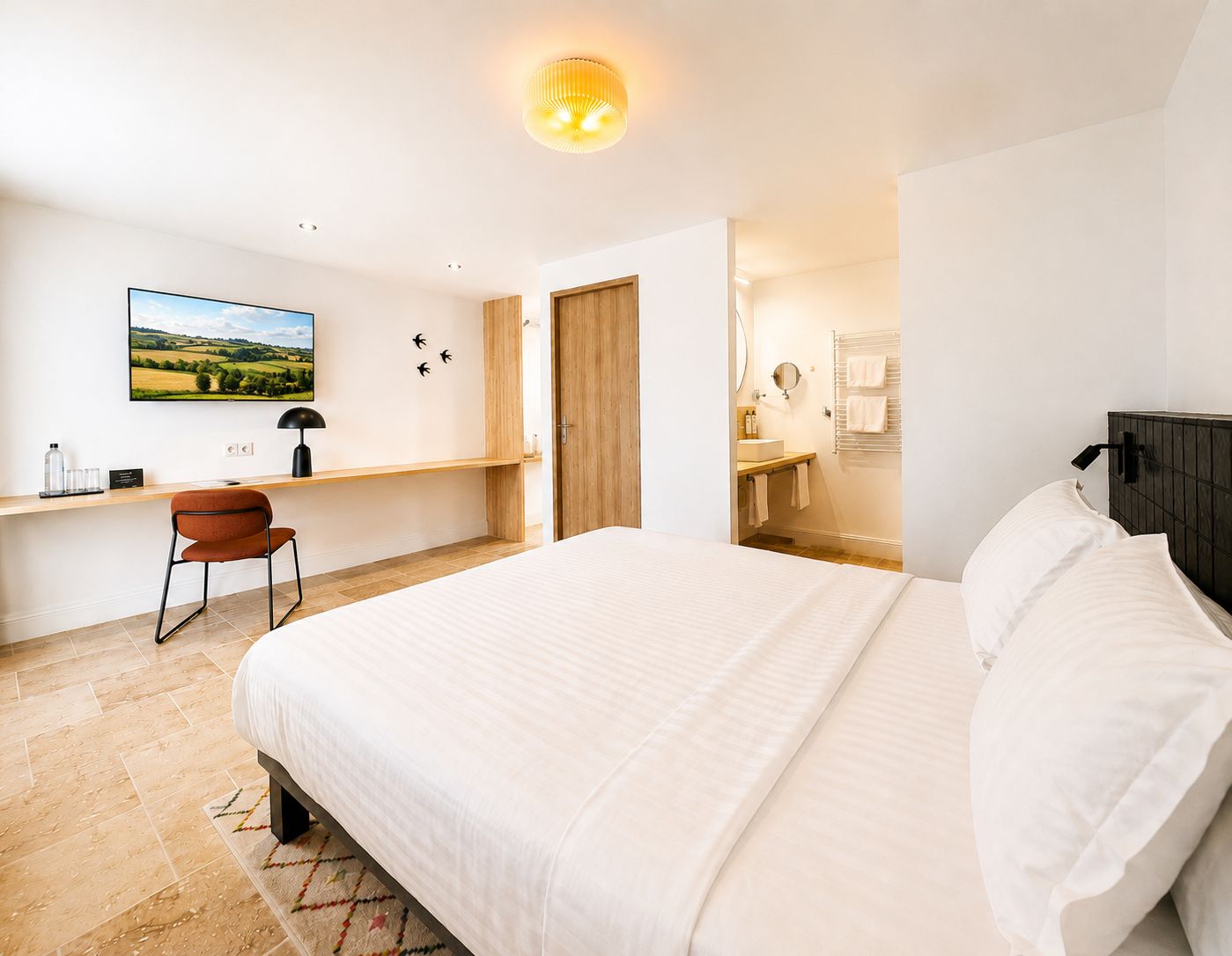 Un dormitorio con una cama grande, un escritorio con silla, un baño con espejo y barra para toallas, y un televisor de pared que muestra un paisaje.