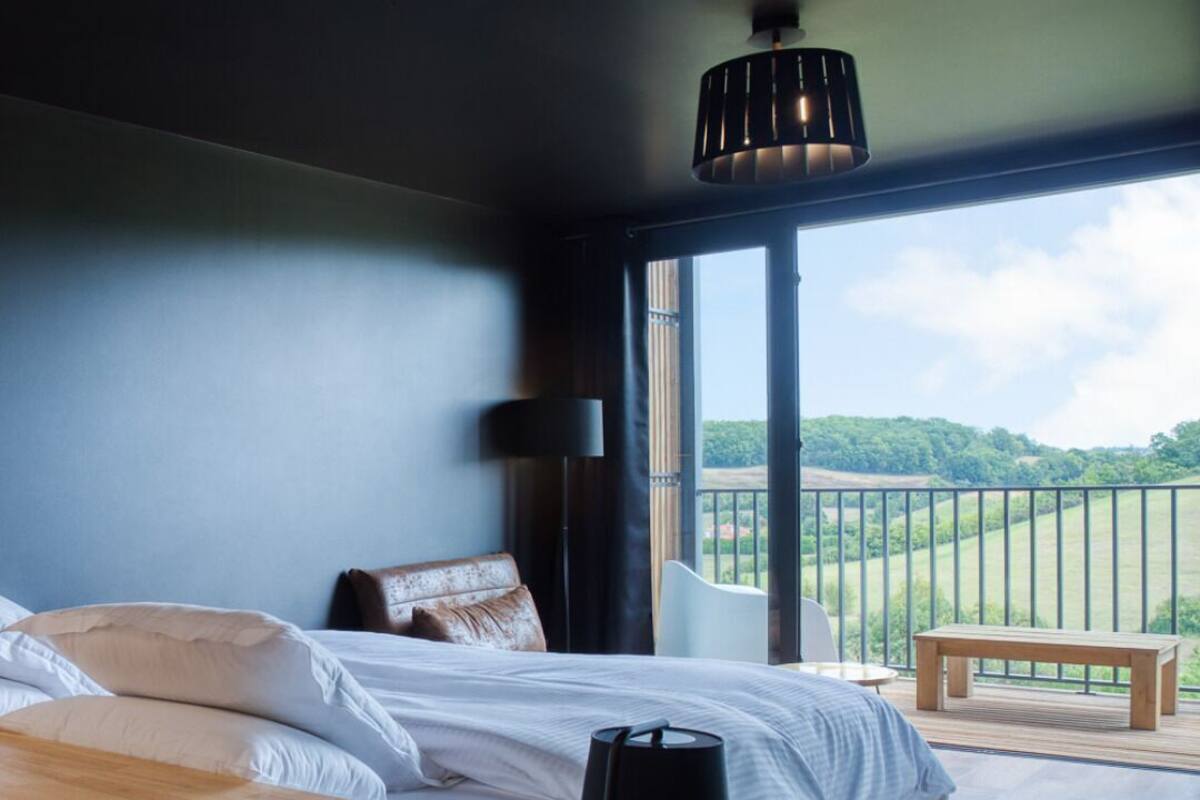 Une chambre avec un grand lit, une table de chevet, une lampe et une vue sur un balcon agrémenté d’un banc en bois.