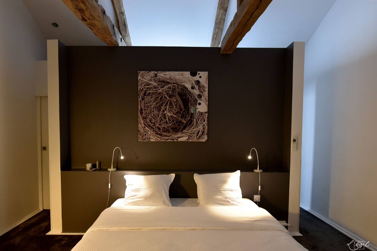 Une chambre à coucher moderne avec un grand lit, deux lampes de chevet, un mur de couleur sombre et une œuvre d’art originale au-dessus du lit.
