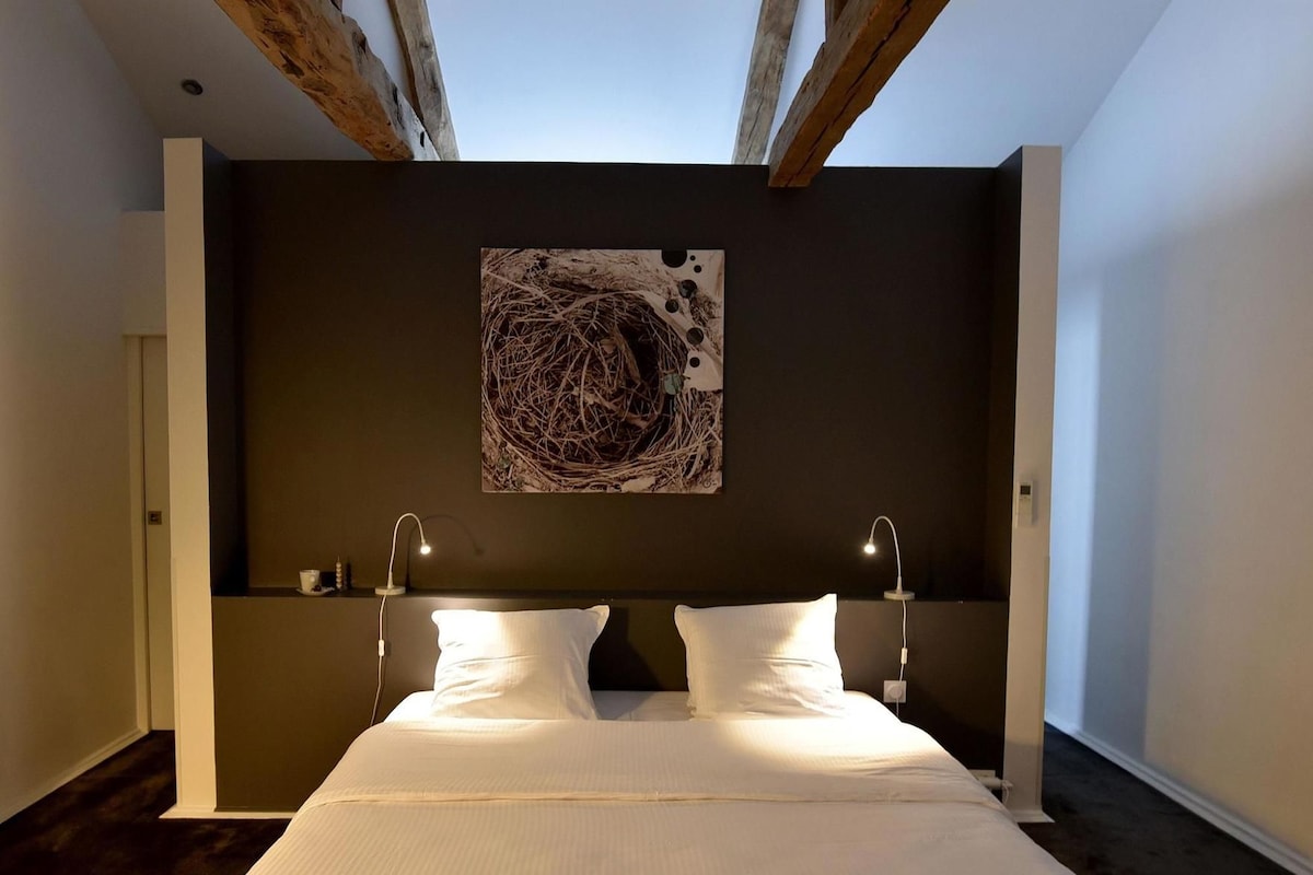 Un dormitorio con una cama grande, dos lámparas de noche, una pared oscura y un techo con vigas a la vista.