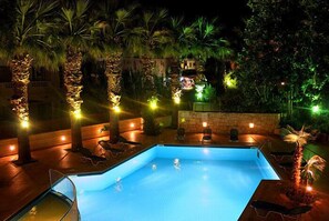 An outdoor swimming pool with lounge chairs and palm trees, surrounded by stone walls and warm lighting. - Latania Apartments (Hersonissos)