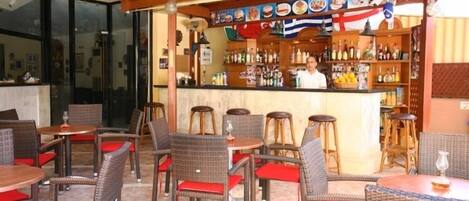 An outdoor bar area with wicker furniture and red cushions, a counter with a bartender, and a variety of bottles and glasses.