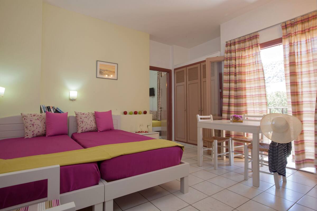 A bedroom with a large bed, a desk, and a chair. There is a wardrobe and a window with curtains.