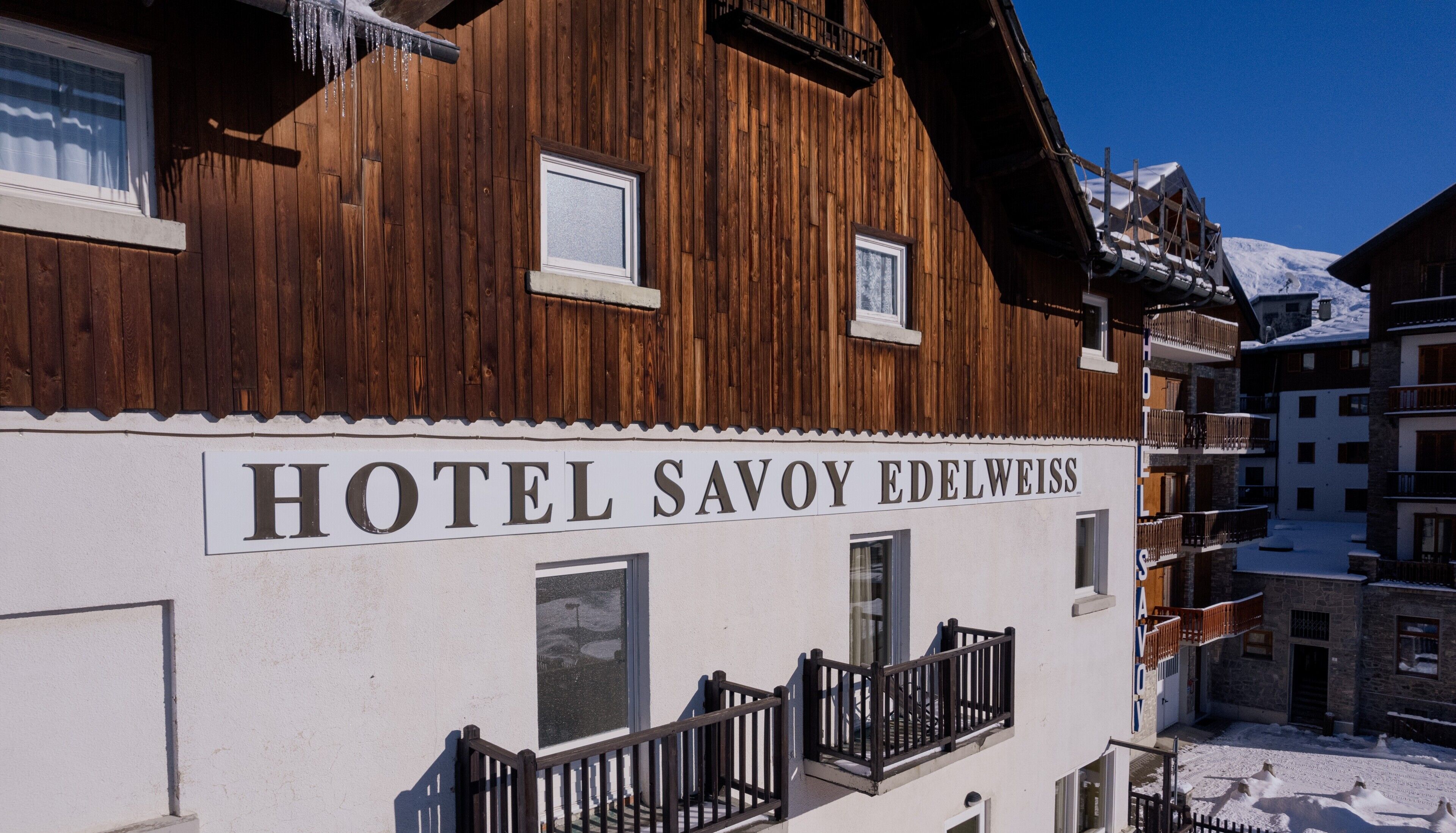 Un cartello con la scritta 'HOTEL SAVOY EDELWEISS' su un edificio bianco con rivestimento in legno.