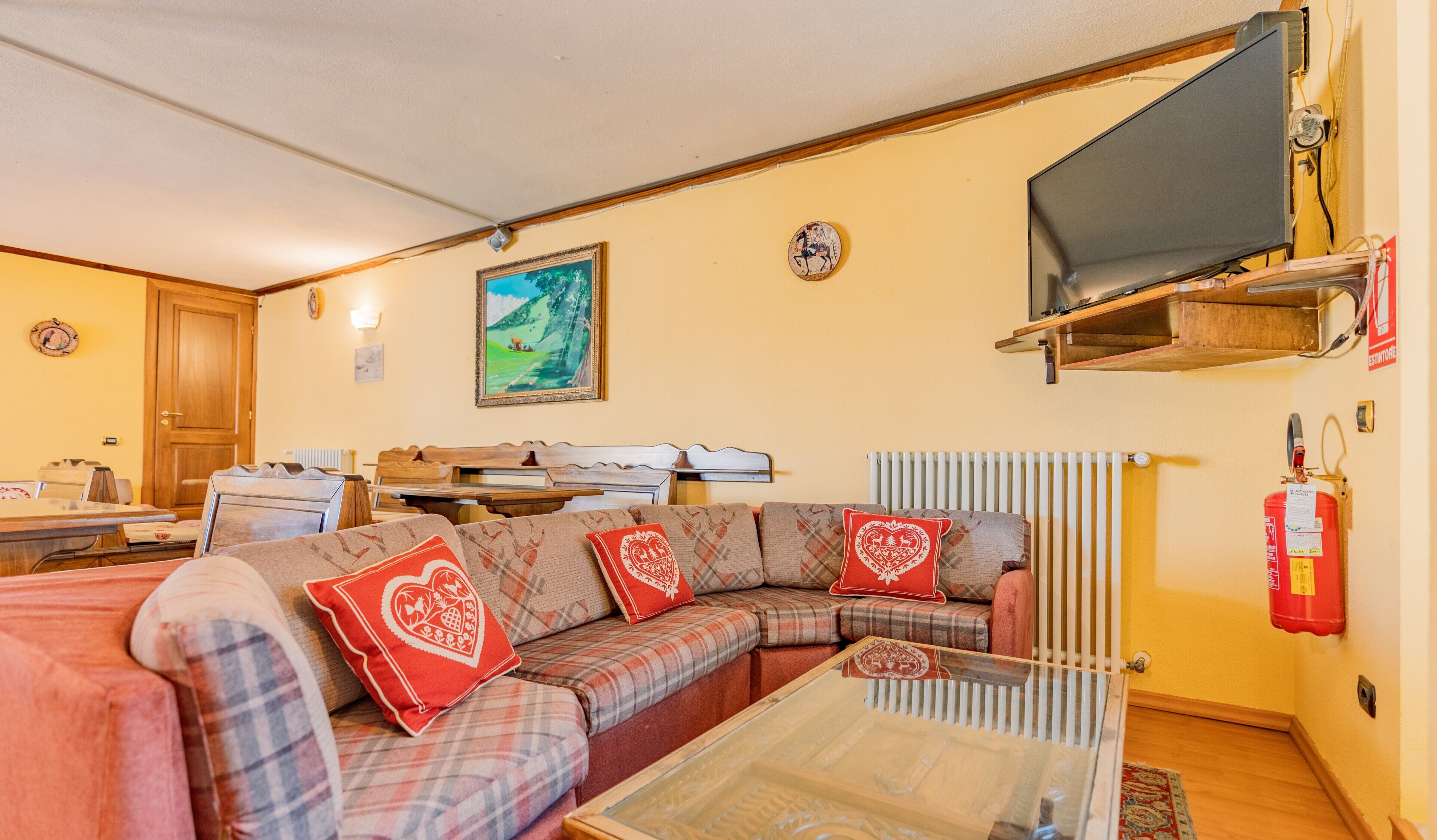 A furnished living room with a sofa set, a television mounted on a wooden shelf, and a fire extinguisher on the wall.