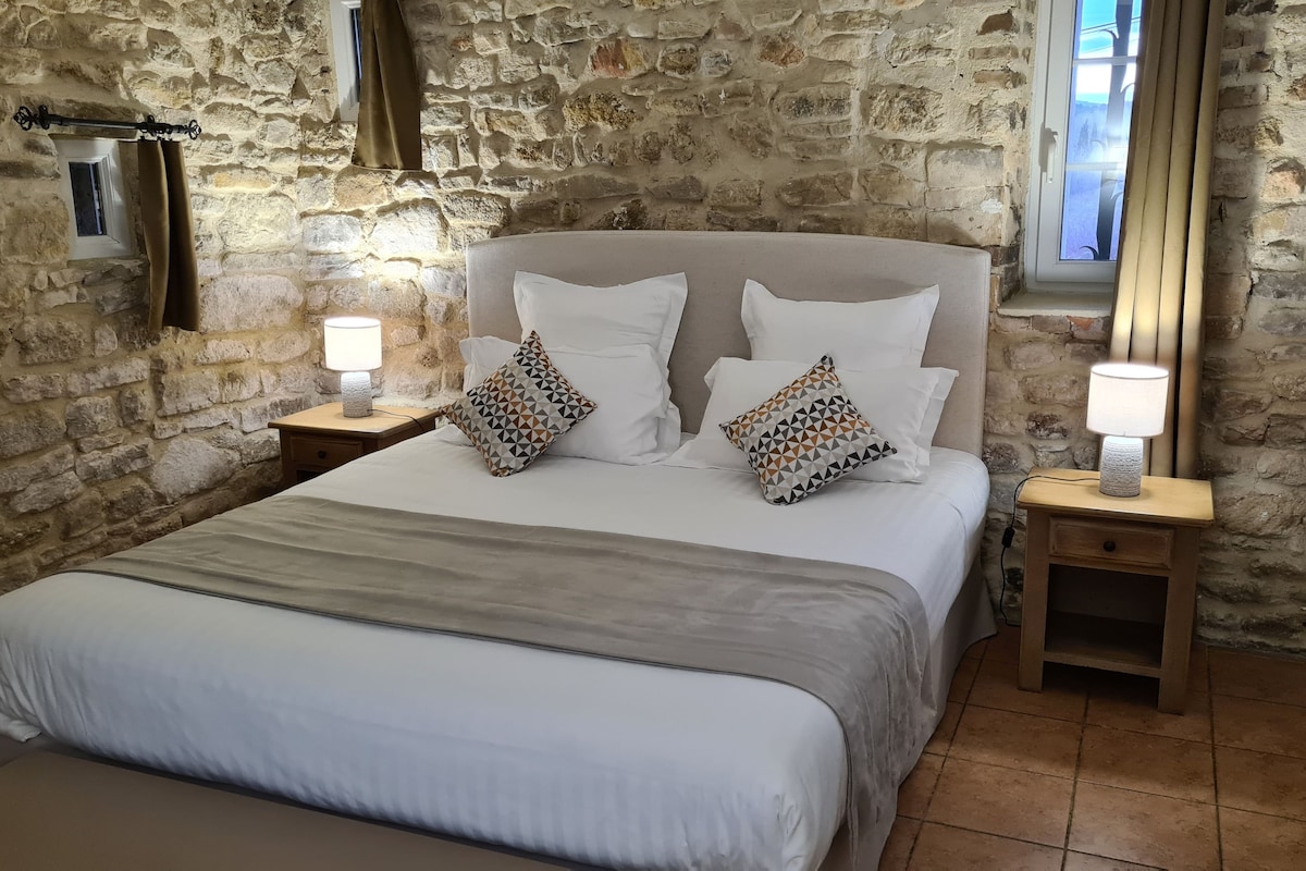 Un dormitorio con pared de piedra, una cama con ropa de cama blanca, dos mesitas de noche con lámparas y una ventana con cortinas.