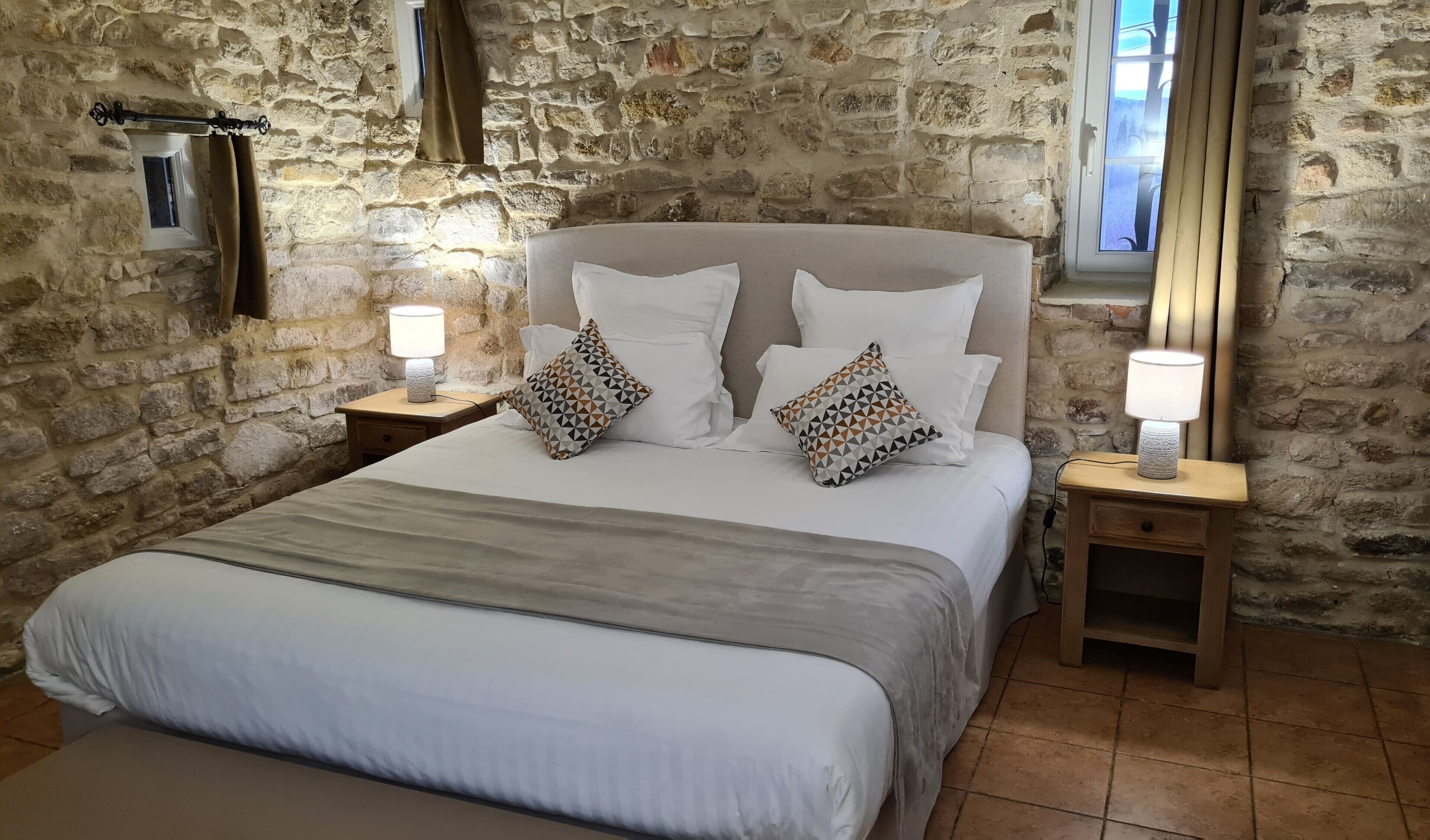 A bedroom with a stone wall, a bed with white bedding, two bedside tables with lamps, and a window with curtains.