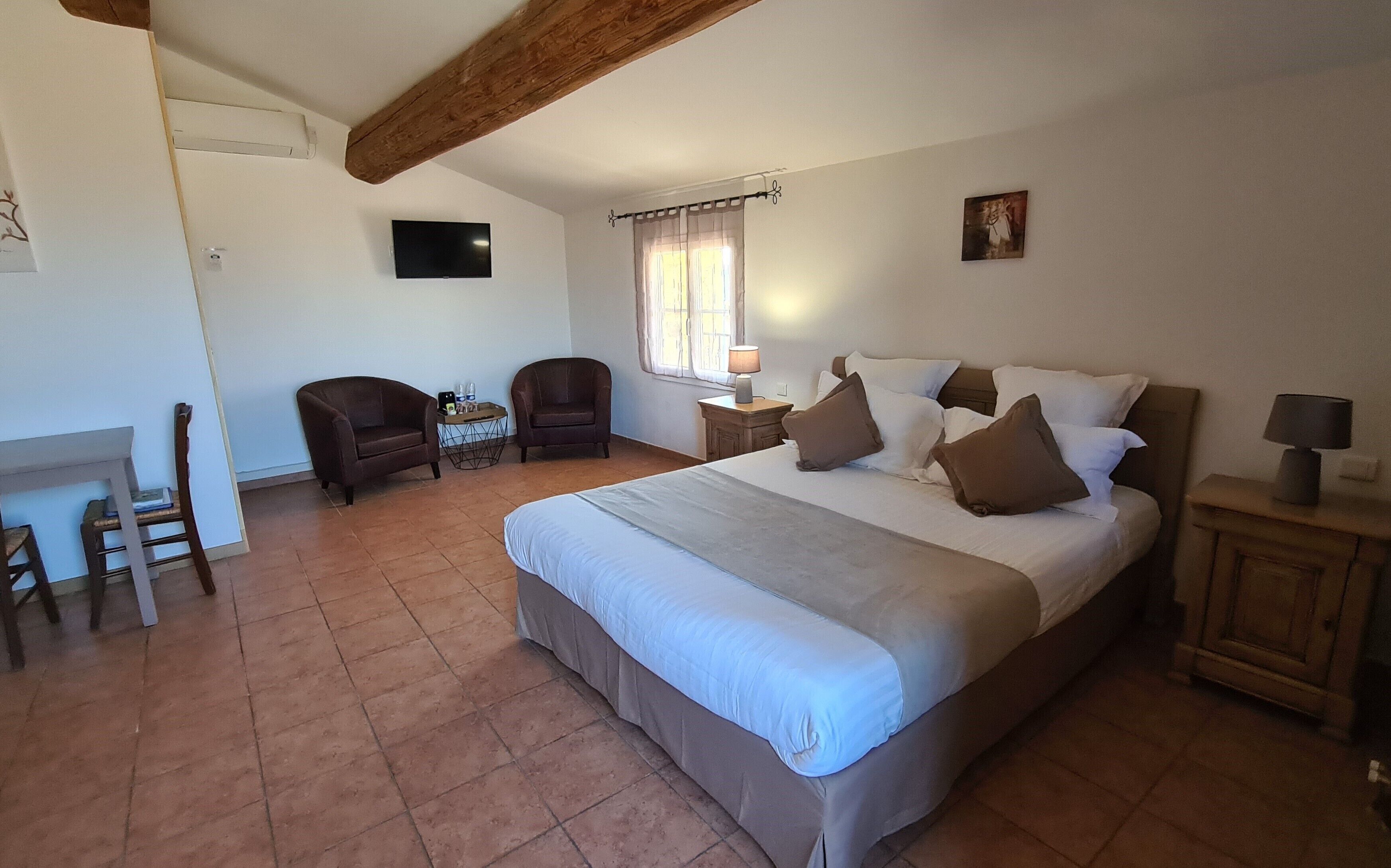A bedroom with a large bed, two armchairs, a small table with bottles, a TV on the wall, and a window with curtains.