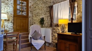 A cozy room with a stone wall, a wooden door, a chair with a blanket, a table with a lamp, and a TV on a wooden cabinet. - Logis Hotel La Bastide St Bach (Suze-la-Rousse)