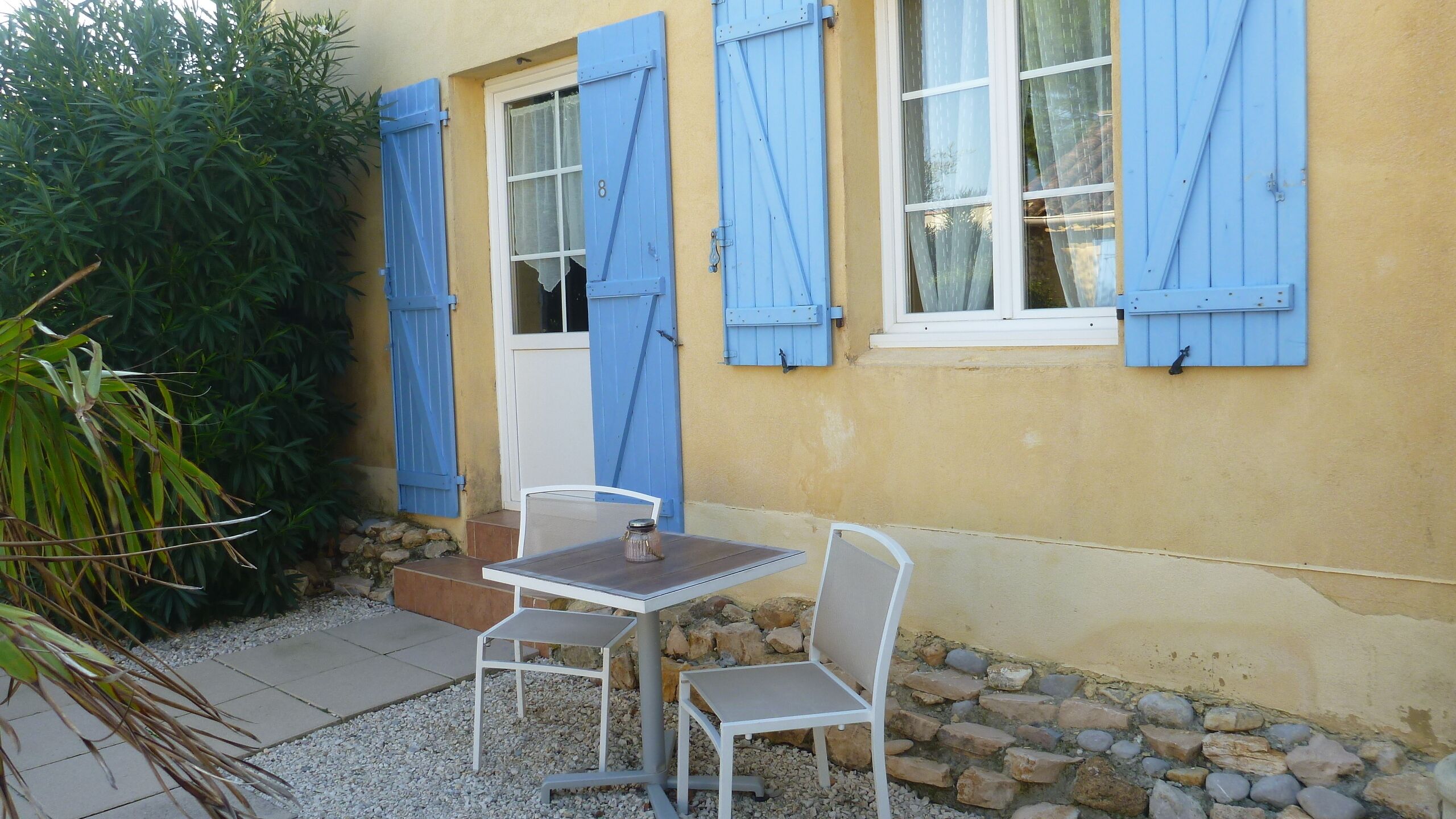 Un pequeño espacio al aire libre con una mesa y dos sillas, una puerta y un ventanal con contraventanas azules.