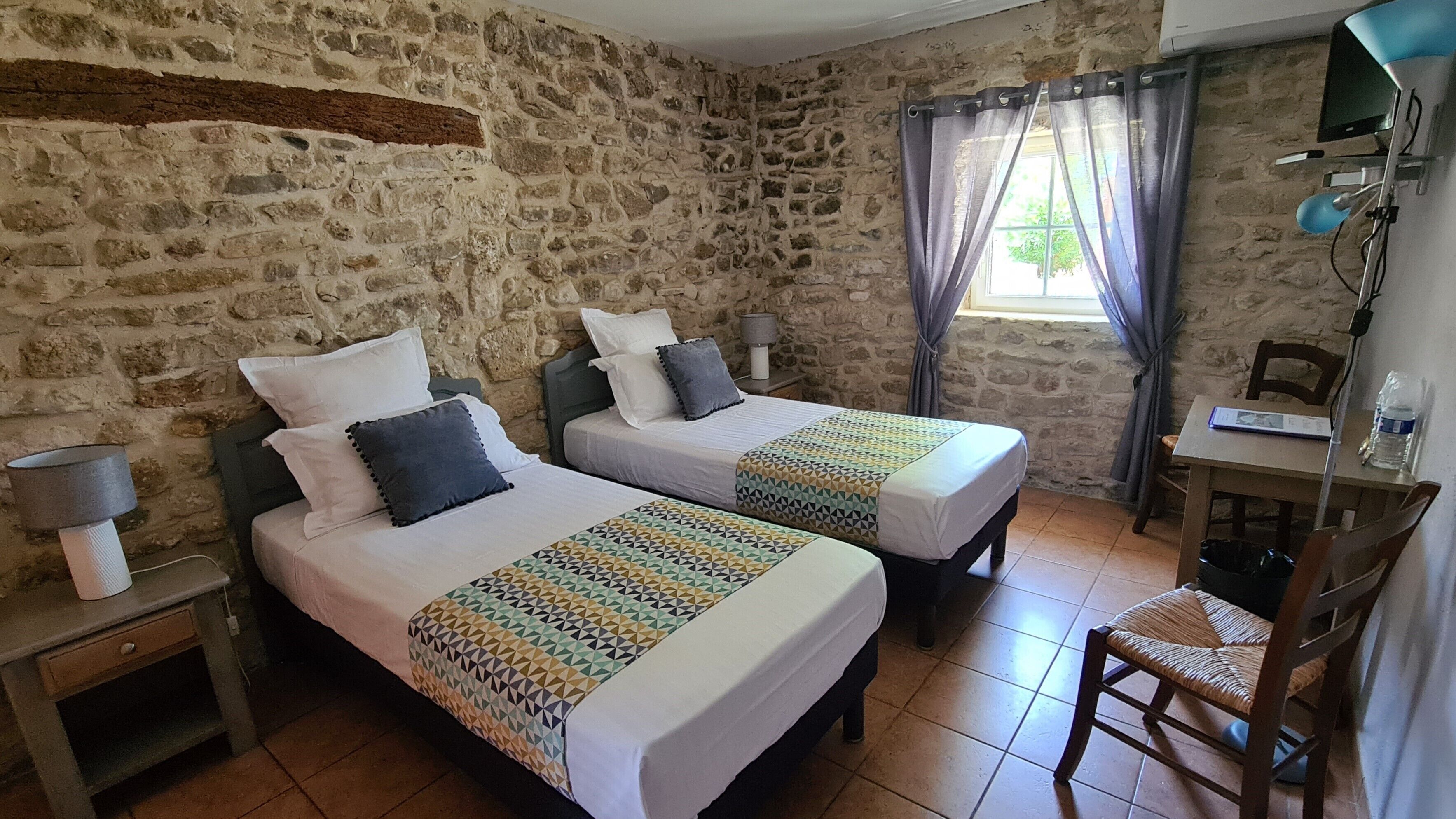 Une chambre avec deux lits, un bureau, une chaise, une lampe et une fenêtre avec des rideaux.