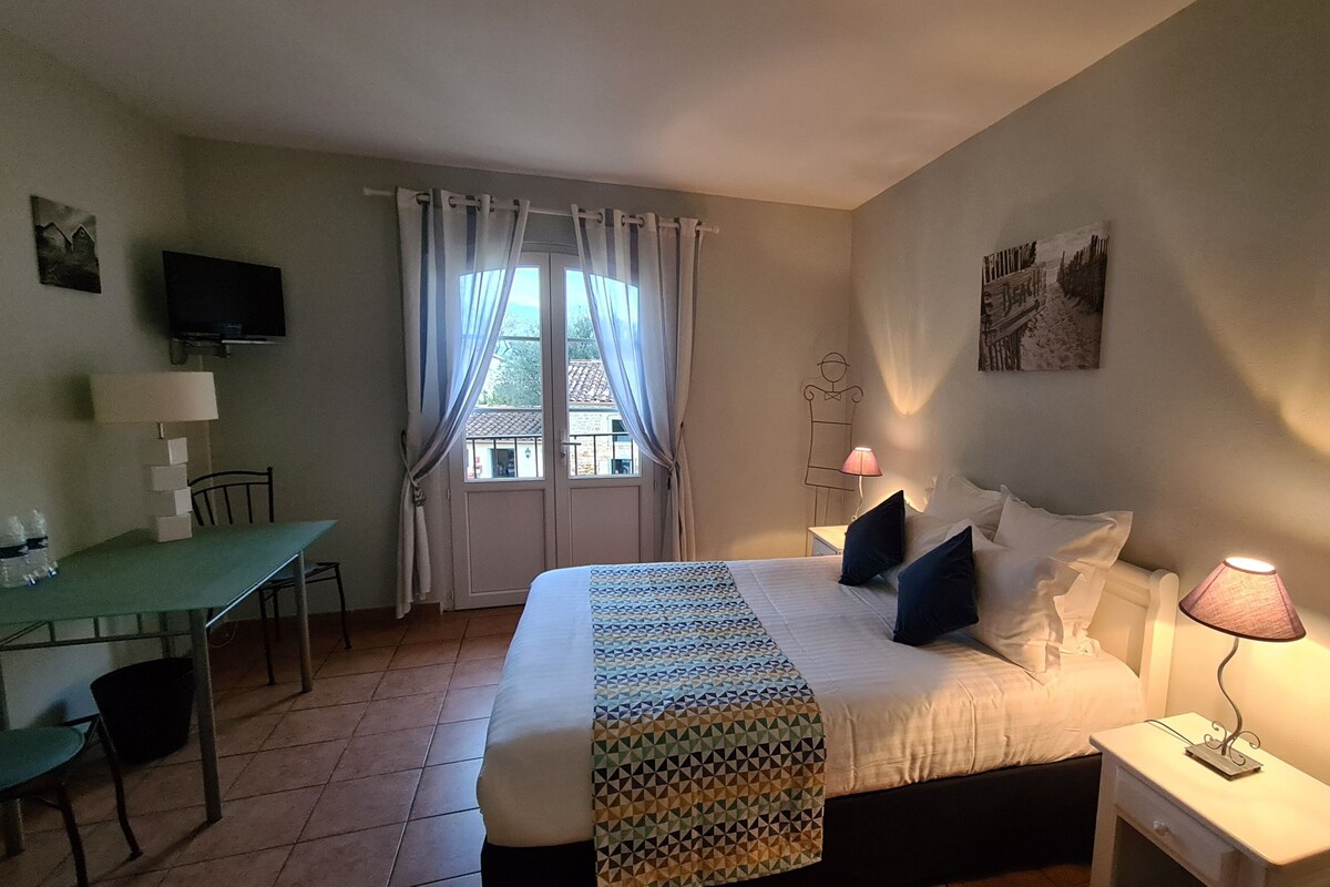 Quarto com cama, mesa de cabeceira, luminária, janela com cortinas, escrivaninha, cadeira e televisão.