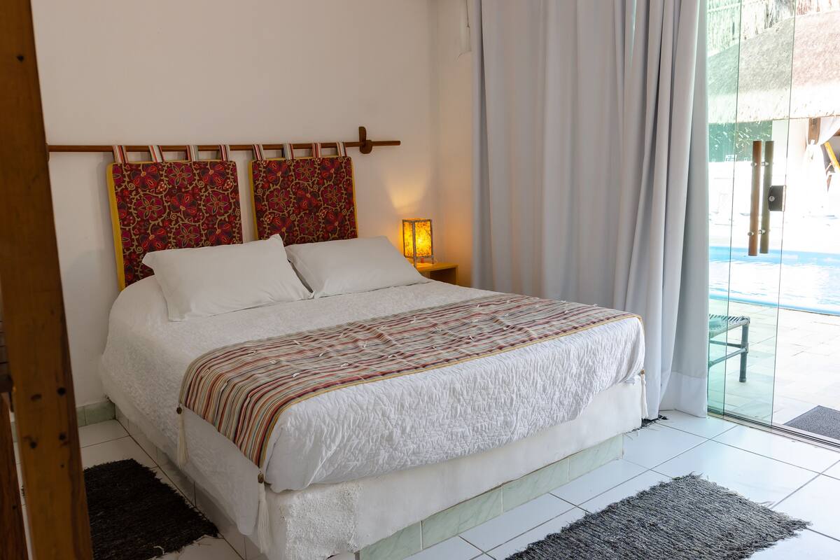 Una cama bien hecha con una colcha estampada, una lámpara de noche y vistas al exterior a través de un ventanal.