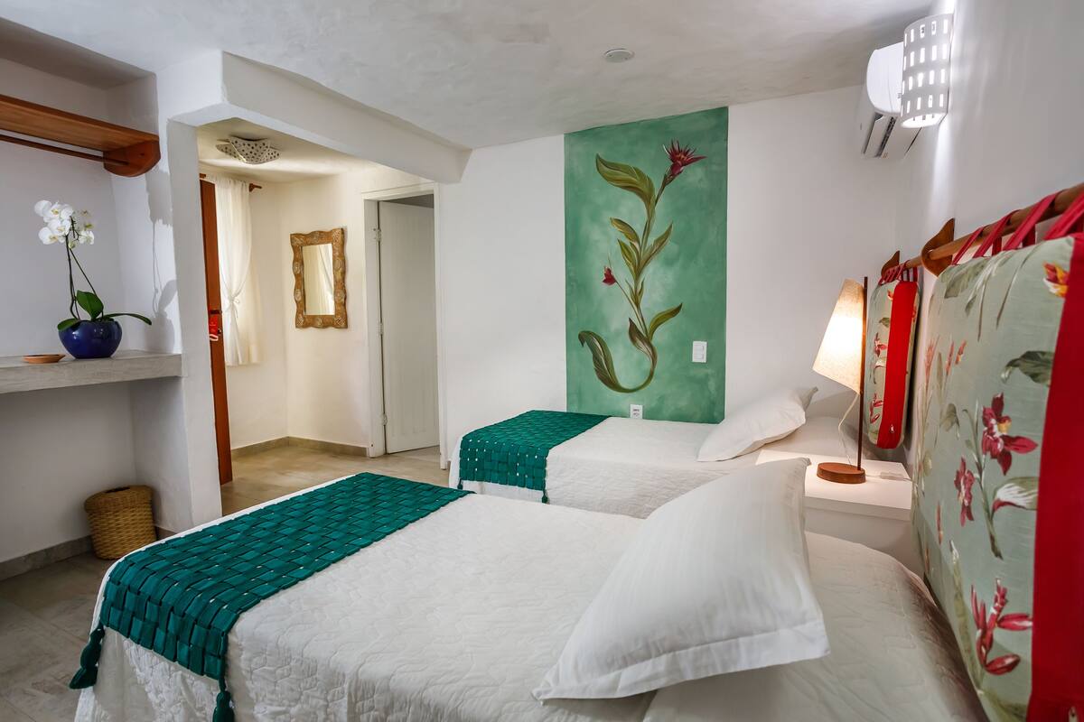 Habitación de hotel con dos camas, un cuadro en la pared color verde, un espejo, una lámpara y una cortina con motivos florales.