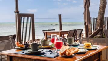 A breakfast table with tropical fruit, juice, and pastries, overlooking a beach.