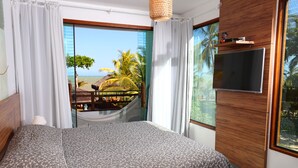 A bedroom with a large bed, a TV mounted on the wall, and a view of the beach and palm trees through the window.