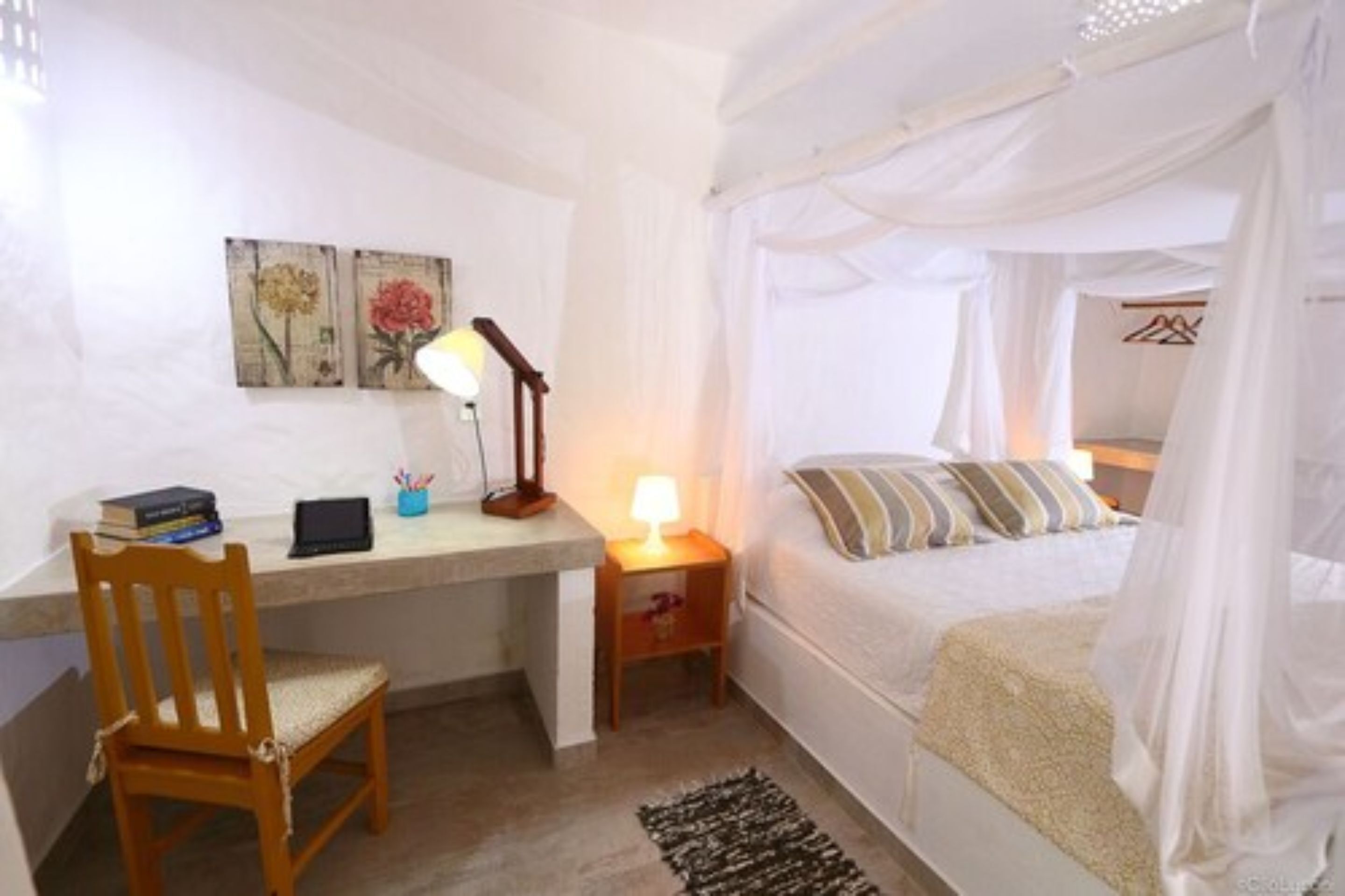 A bedroom with a bed under a canopy, a desk with a chair, a bedside table with a lamp, and two framed pictures on the wall.