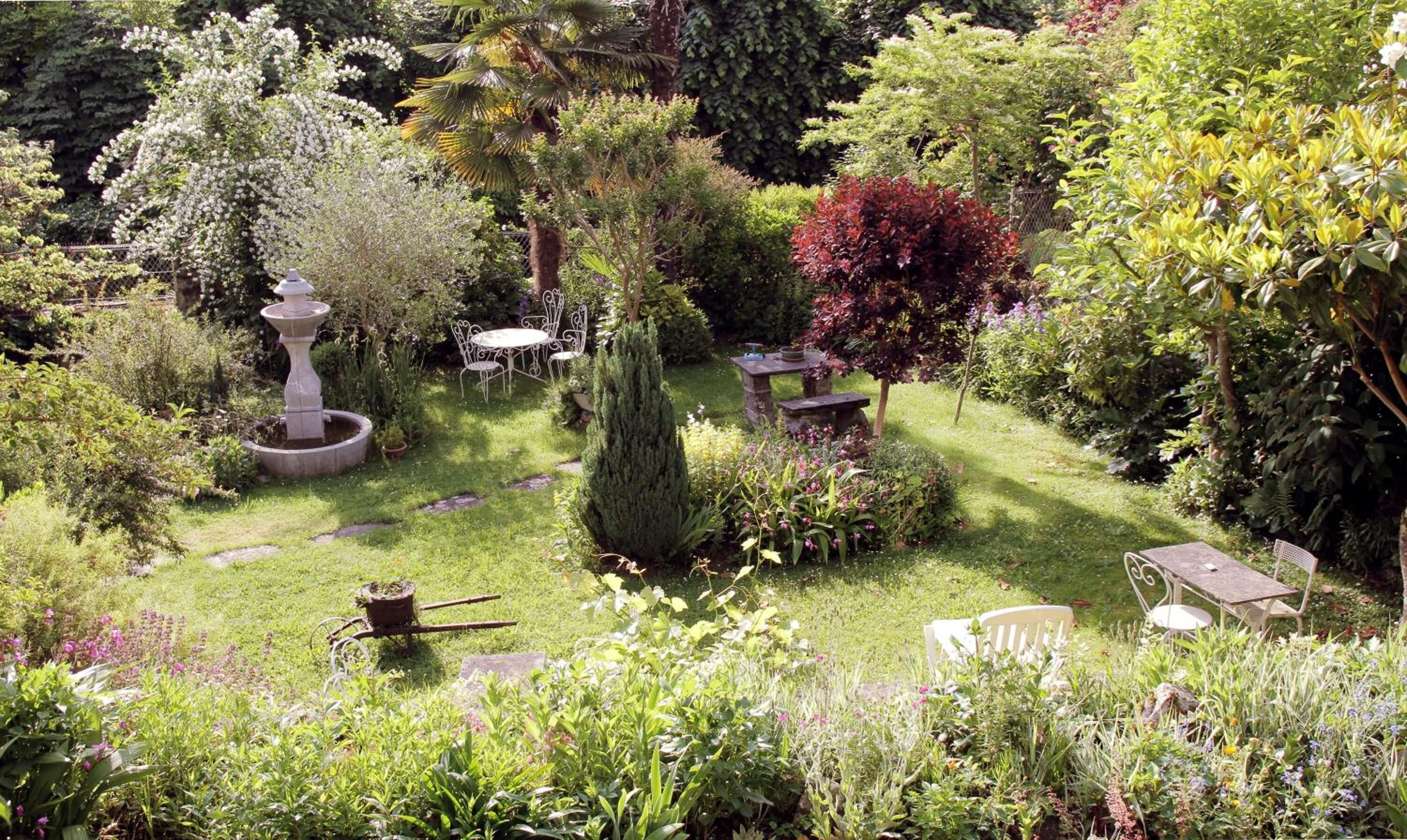 A lush garden with a variety of plants, a stone fountain, and outdoor seating.