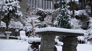 Un banco de piedra en un jardín nevado, con un edificio al fondo.