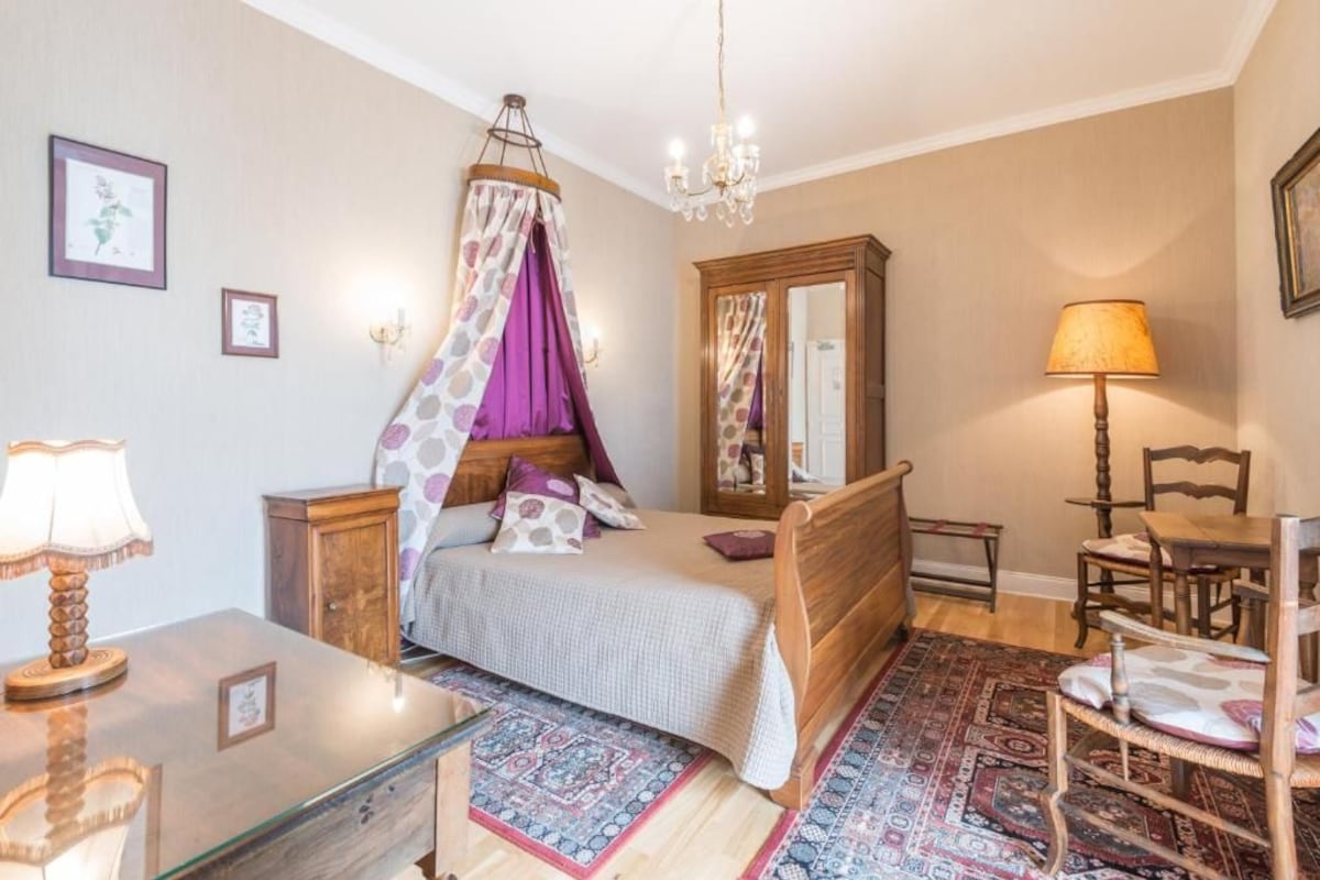 Une chambre à coucher comprenant un lit, une table de chevet en bois, une table en verre, une chaise, une lampe, un lustre, un miroir et un tapis à motifs.