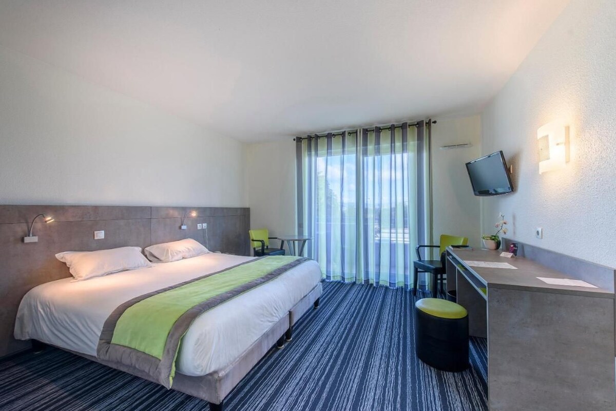 Habitación de hotel con cama, escritorio, silla, televisor y ventana con cortinas.