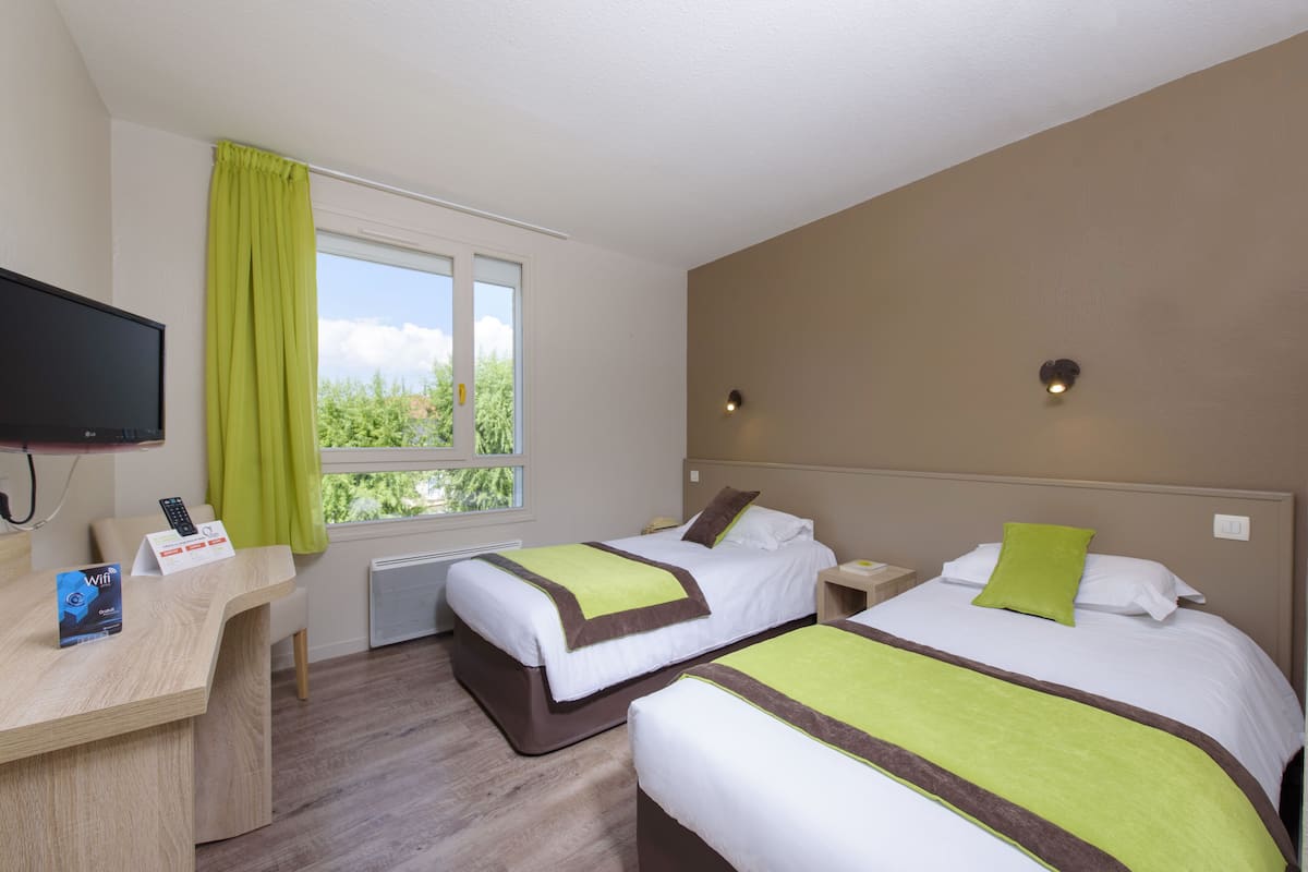 A hotel room with two beds, a desk with a monitor, and a window with a view of greenery.