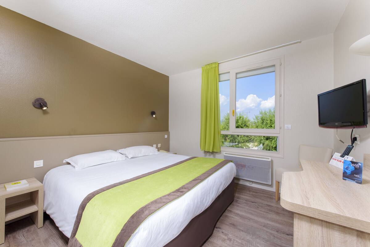 A hotel room with a bed, a desk with a computer, a window with curtains, and a TV.