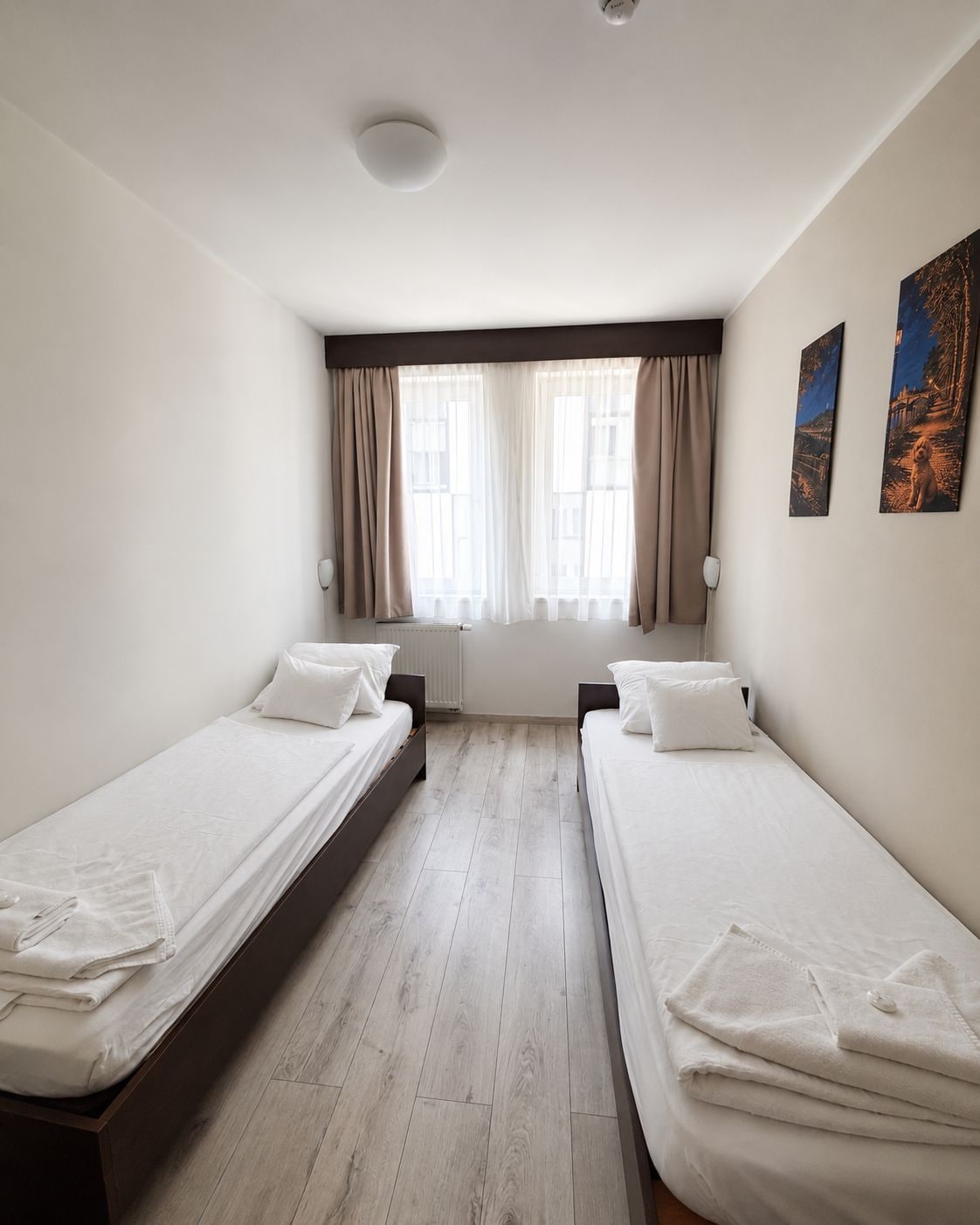 Hotellihuone, jossa on kaksi erillistä sänkyä, patteri, ikkuna verhoilla ja seinään kiinnitetty lamppu.