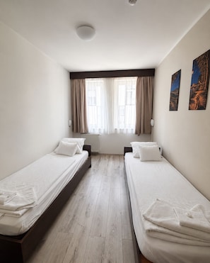 A hotel room with two single beds, a radiator, a window with curtains, and a wall-mounted lamp.