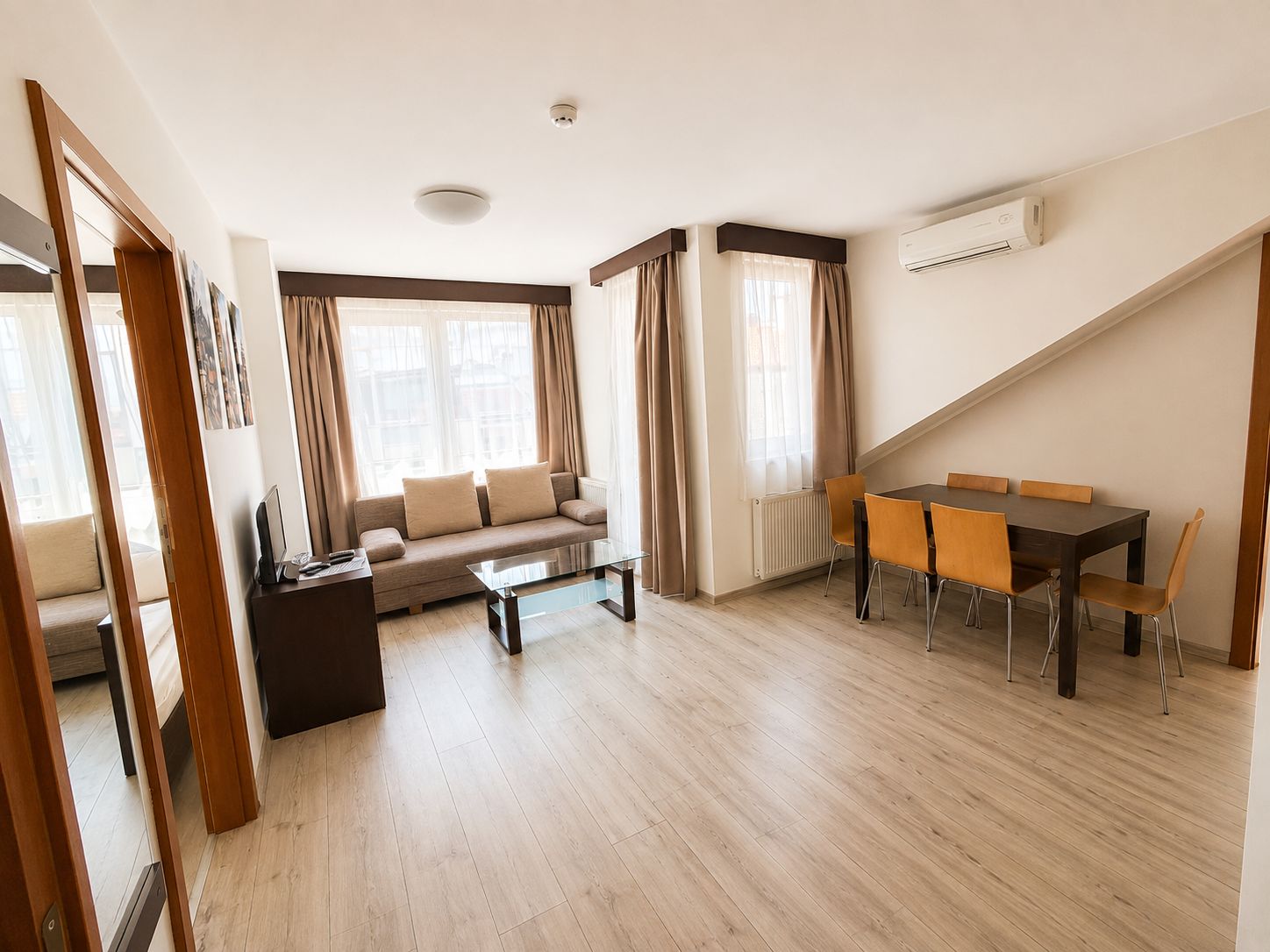Un salon moderne avec un canapé, une table basse et une table à manger avec des chaises. On y trouve une fenêtre avec des rideaux, un climatiseur et un luminaire au plafond.