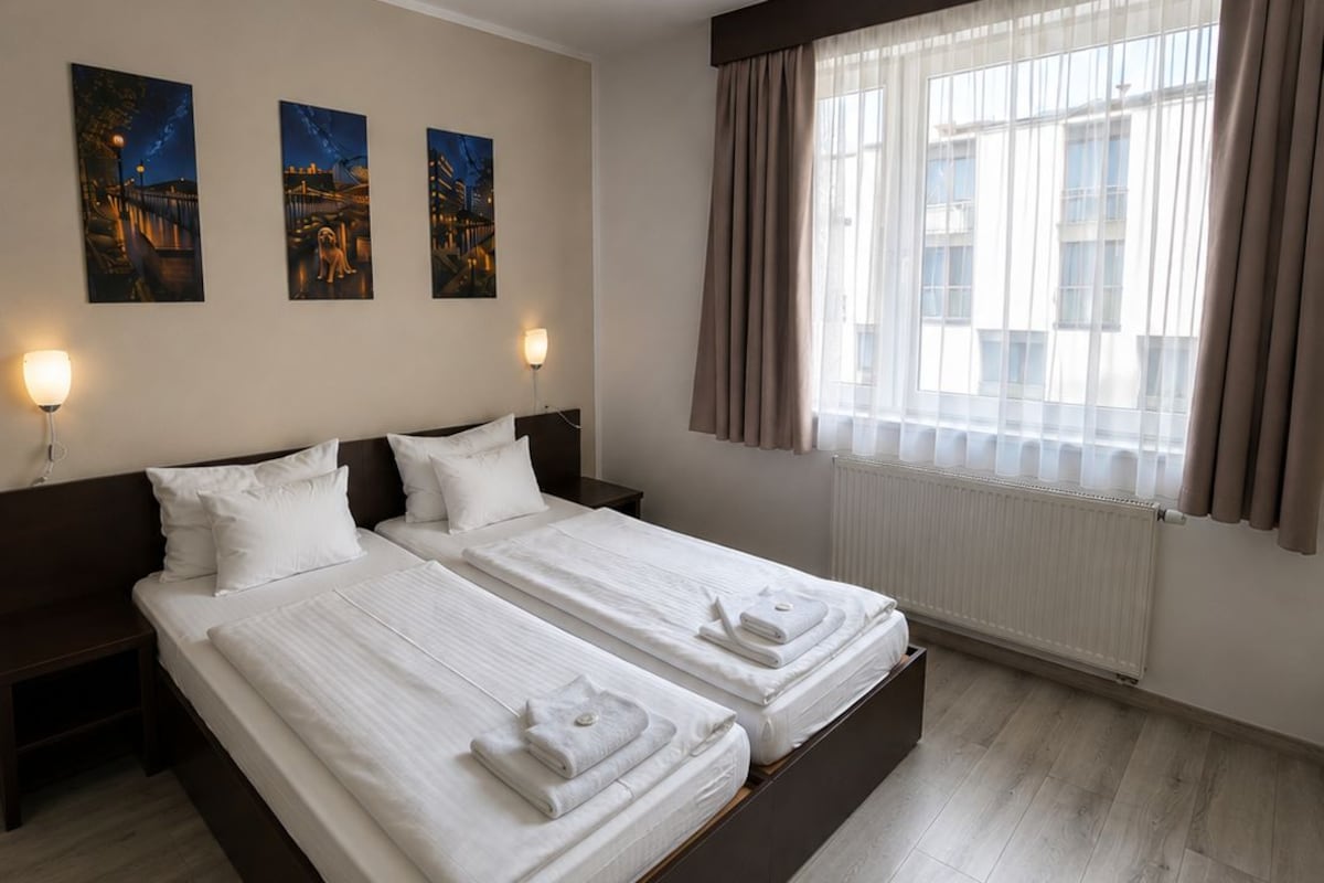 A double bed with white linens and towels, a nightstand, a wall-mounted lamp, and a window with curtains.