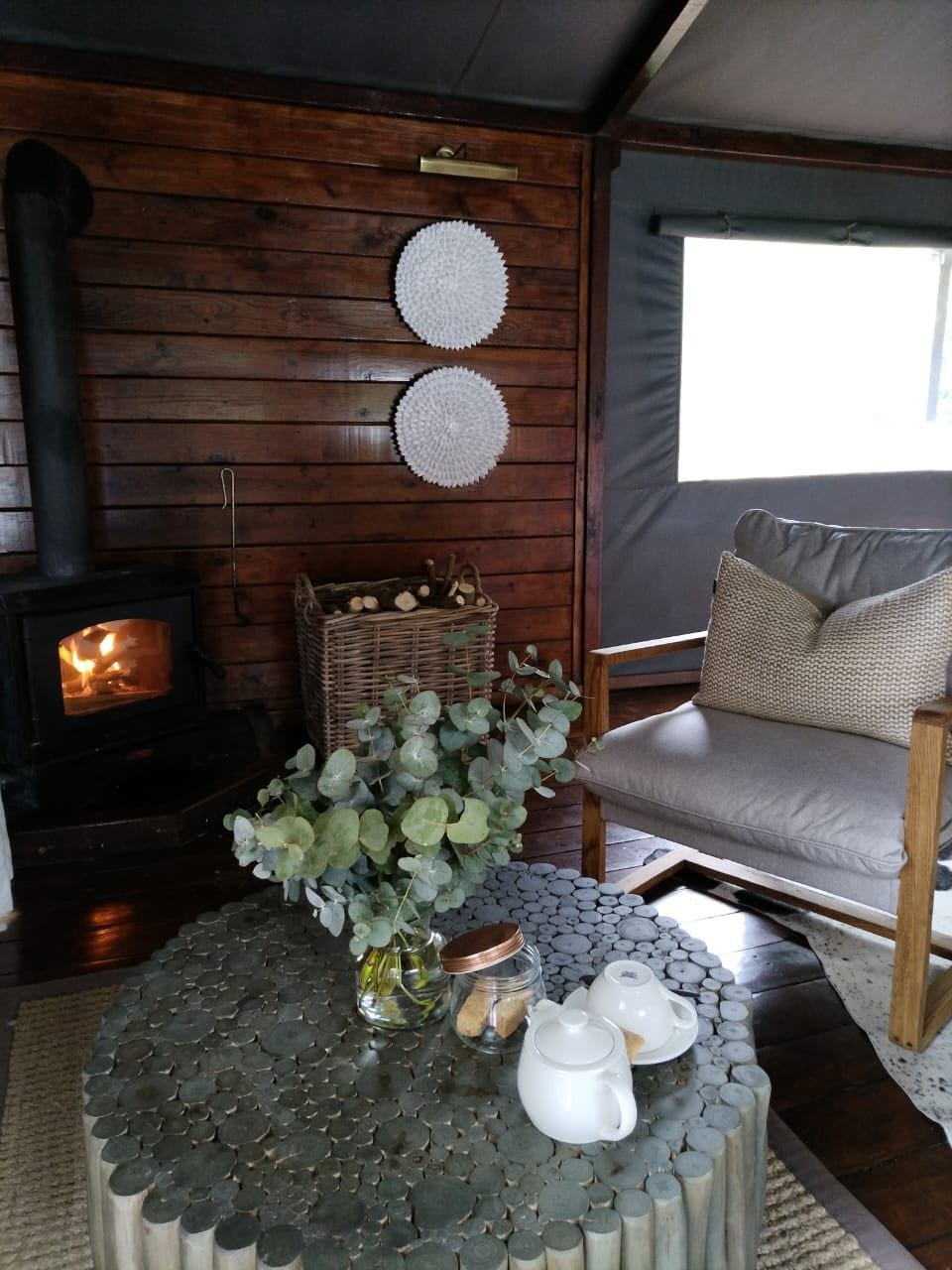 A cozy living room with a wood-burning stove, a wicker basket, a sofa, and a coffee table with a vase and teapot.
