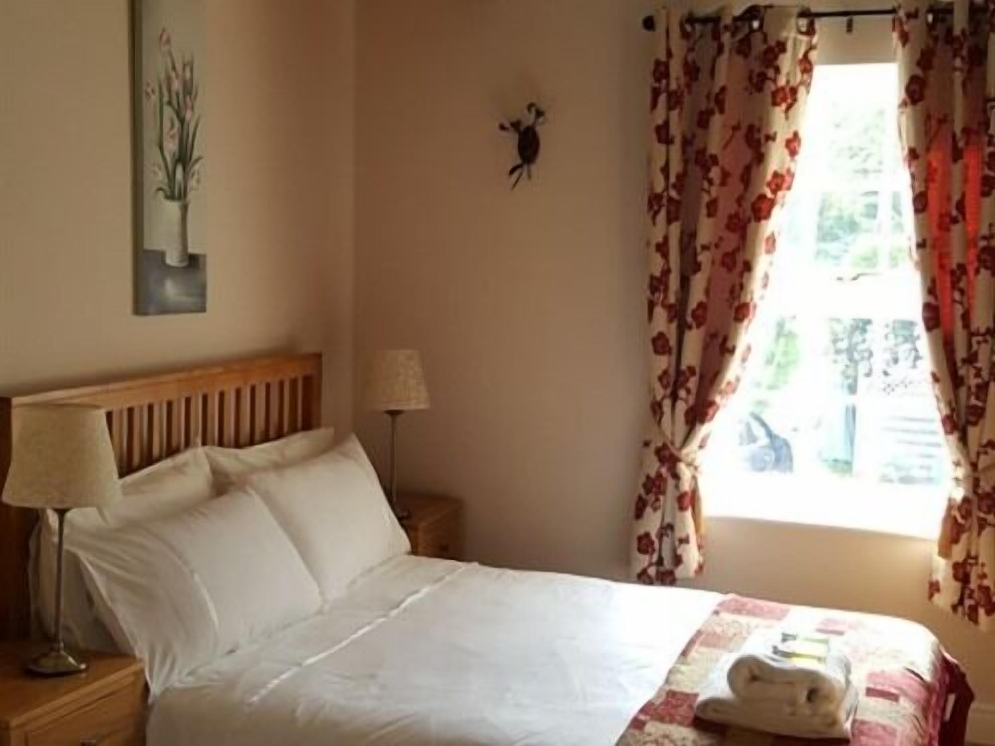 A bedroom with a bed, a nightstand, a lamp, a wall-mounted artwork, and a window with floral curtains.