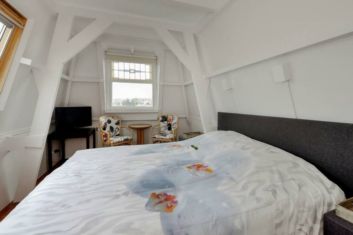 A bedroom with a large bed, two chairs, a small table, a TV, and a window with a city view.