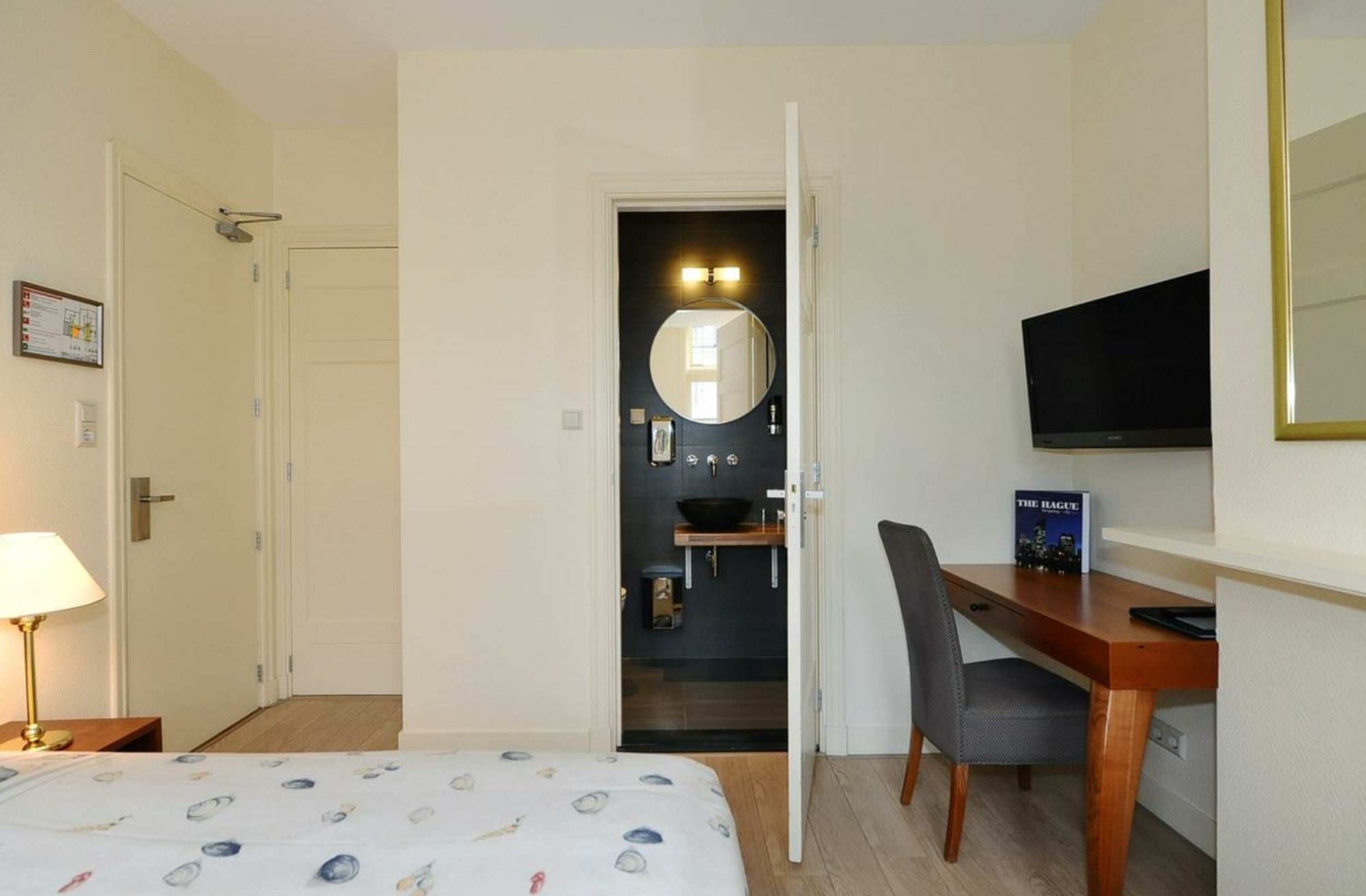 A hotel room with a bed, a desk with a chair, a TV, a mirror, and a bathroom visible through an open door.