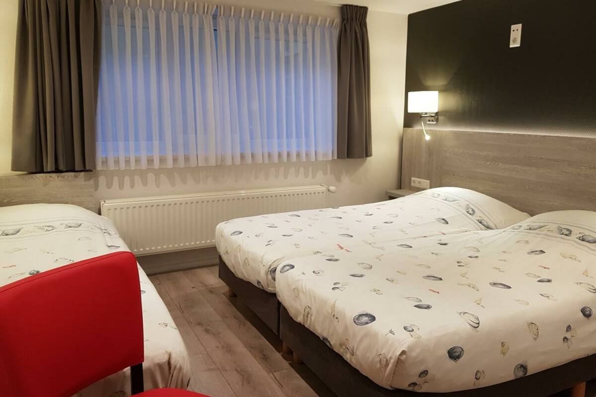 A hotel room with two beds, a red chair, a window with curtains, a wall-mounted lamp, and a radiator.