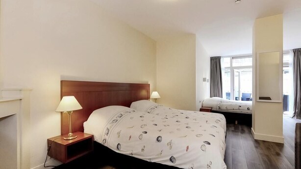 A hotel room with a wooden headboard bed, a bedside table with a lamp, a second bed in the background, and a large window with curtains. - Hotel Bor Scheveningen (The Hague)