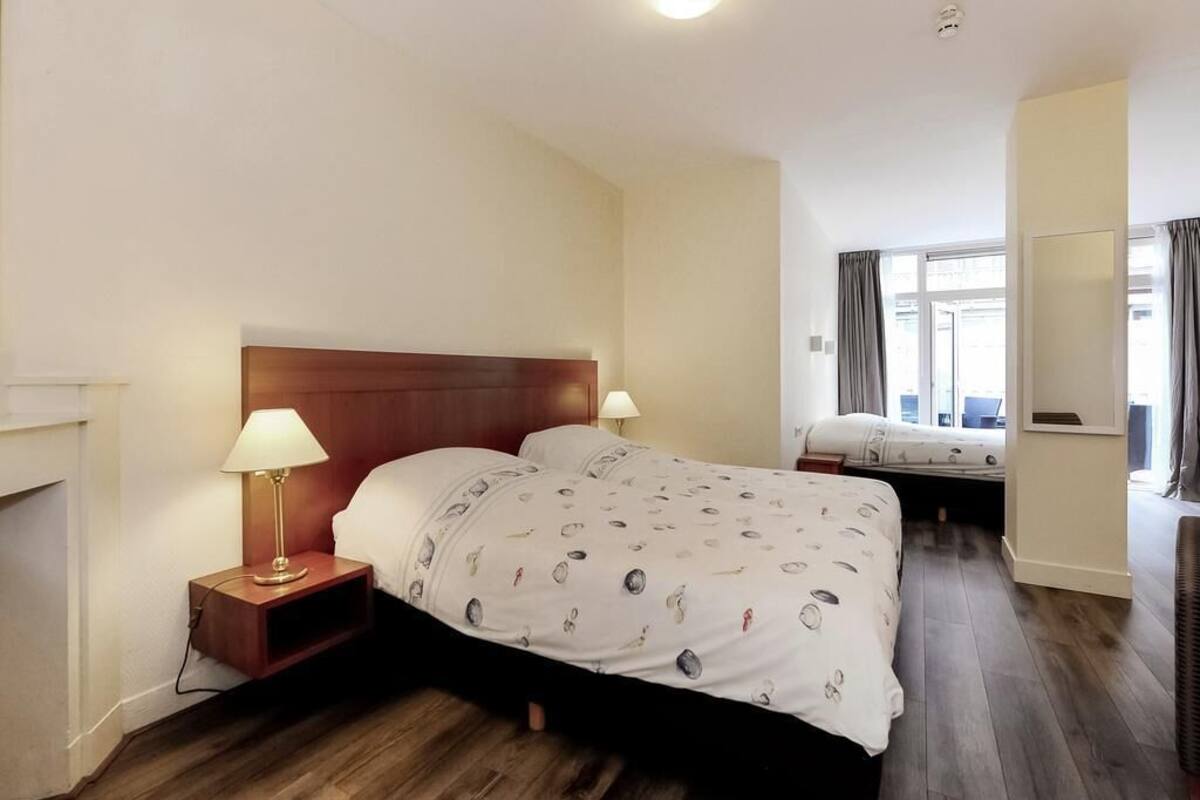A hotel room with a wooden headboard bed, a bedside table with a lamp, a second bed in the background, and a large window with curtains.